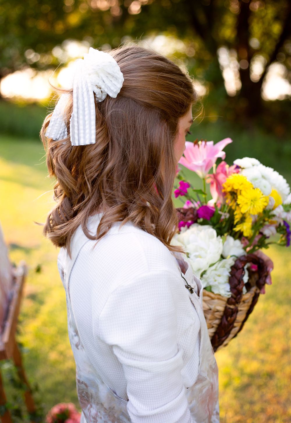 Woman in white with hair bow holds colorful flowers against sunset garden backdrop.