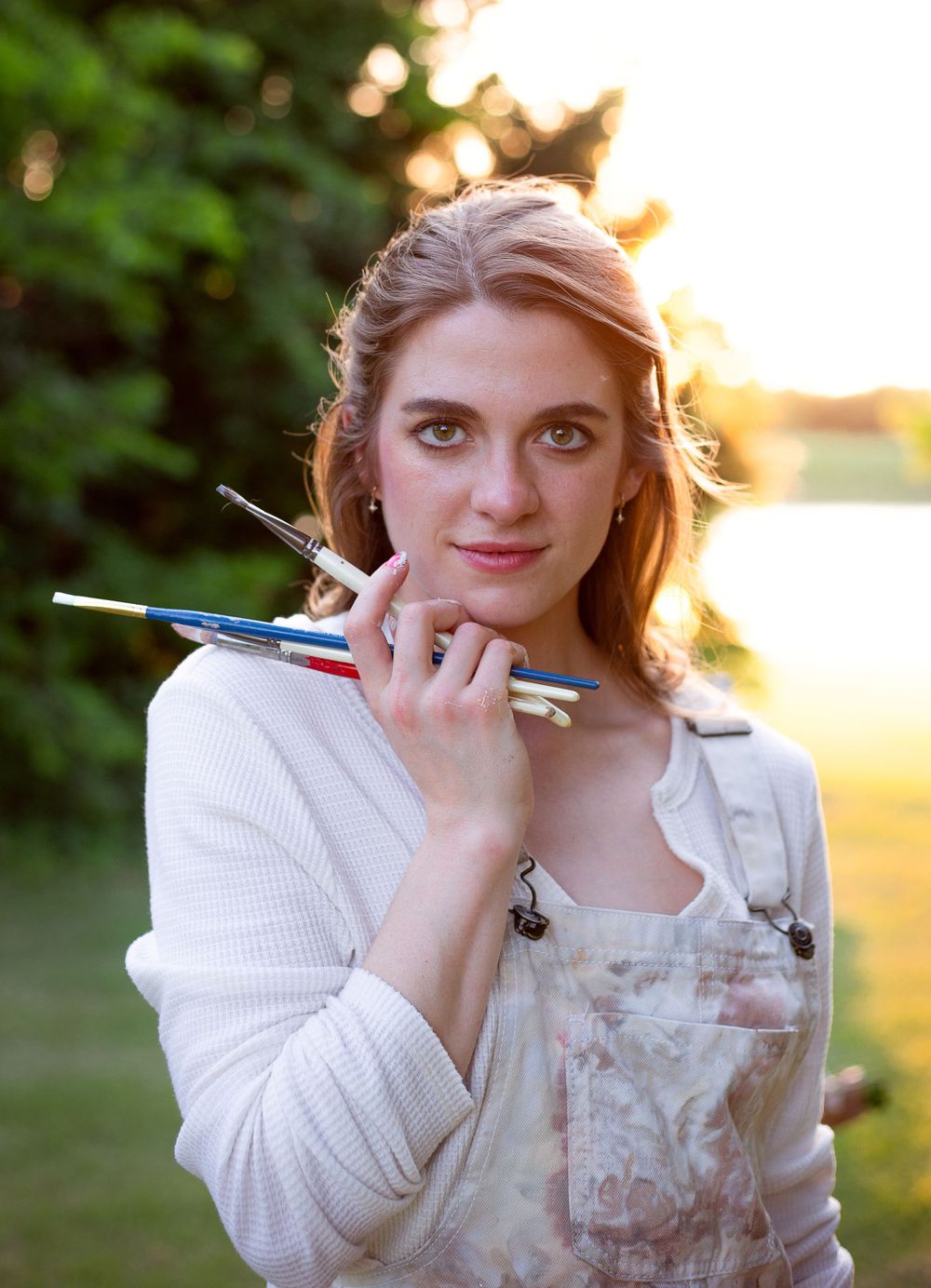 Artist in white sweater holds paintbrushes at golden hour outdoors.