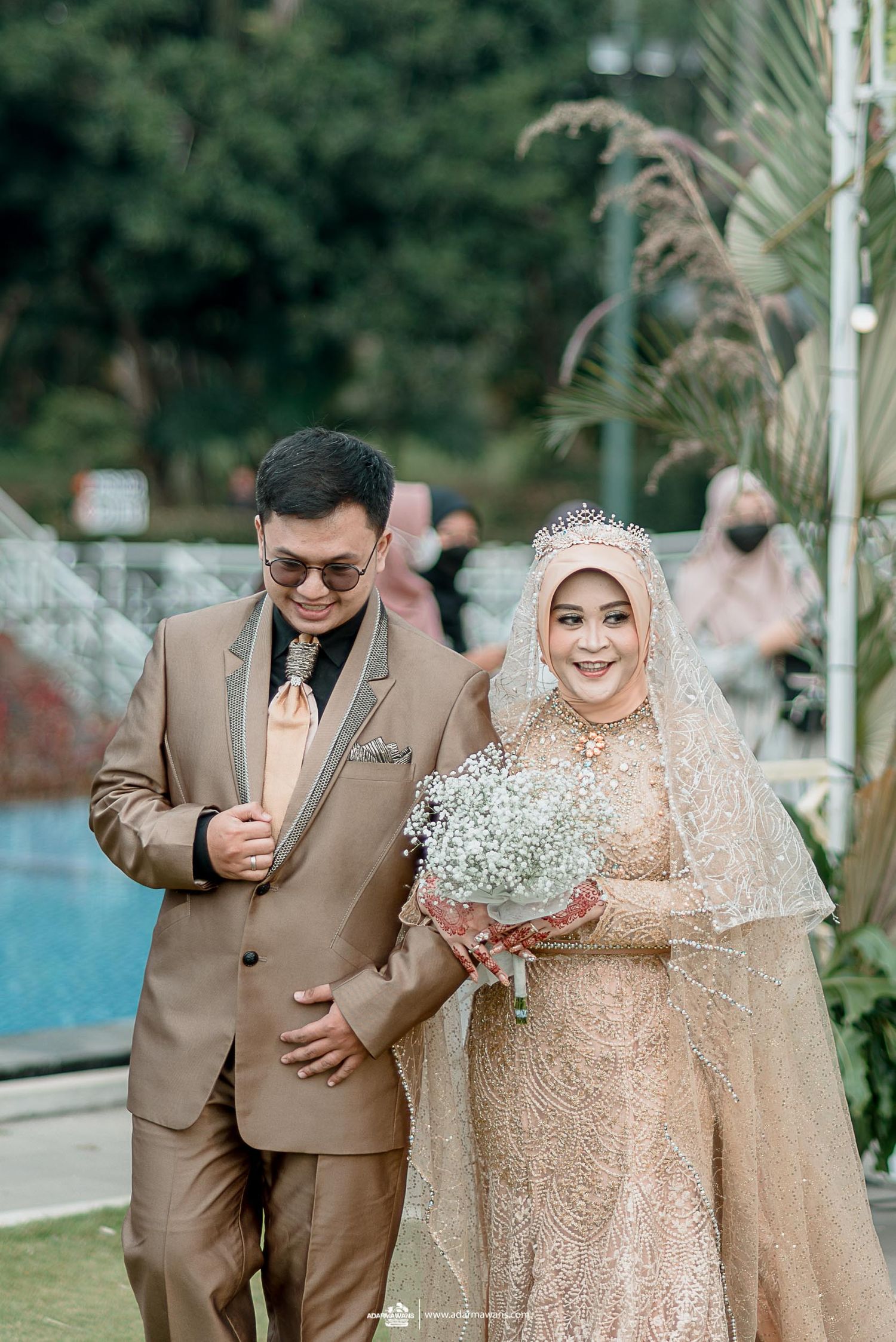 Wedding couple in beige attire pose with baby's breath bouquet in outdoor garden setting.
