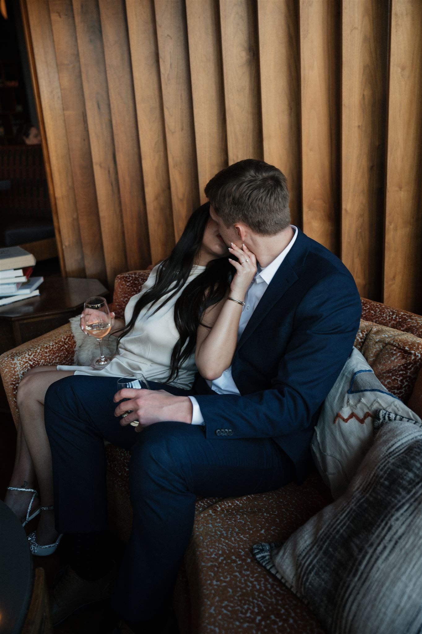 Couple sharing tender moment on brown leather couch with wine glasses in atmospheric lounge.