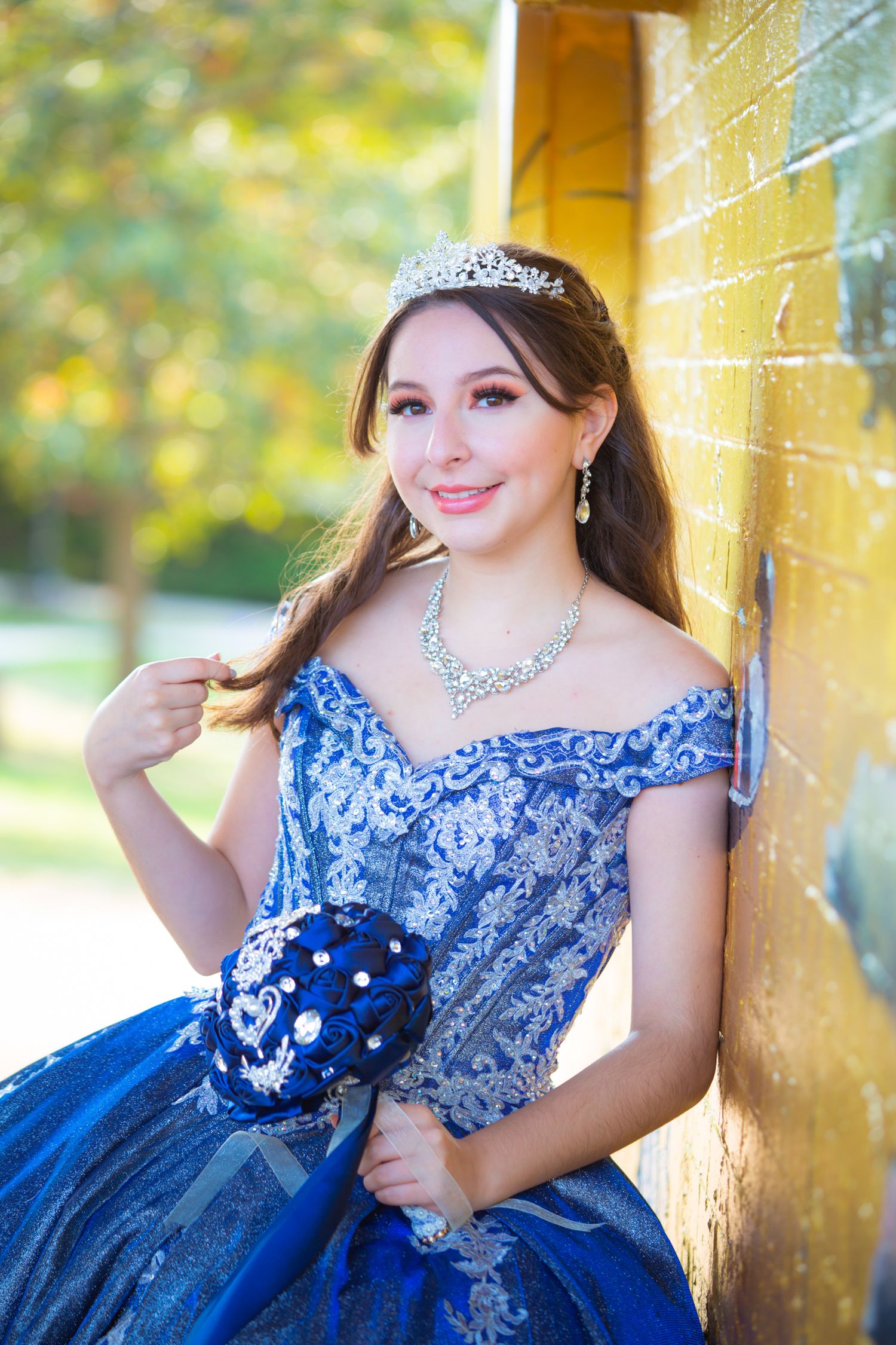 Elegant portrait in a blue ballgown with crystal tiara against a yellow wall backdrop.