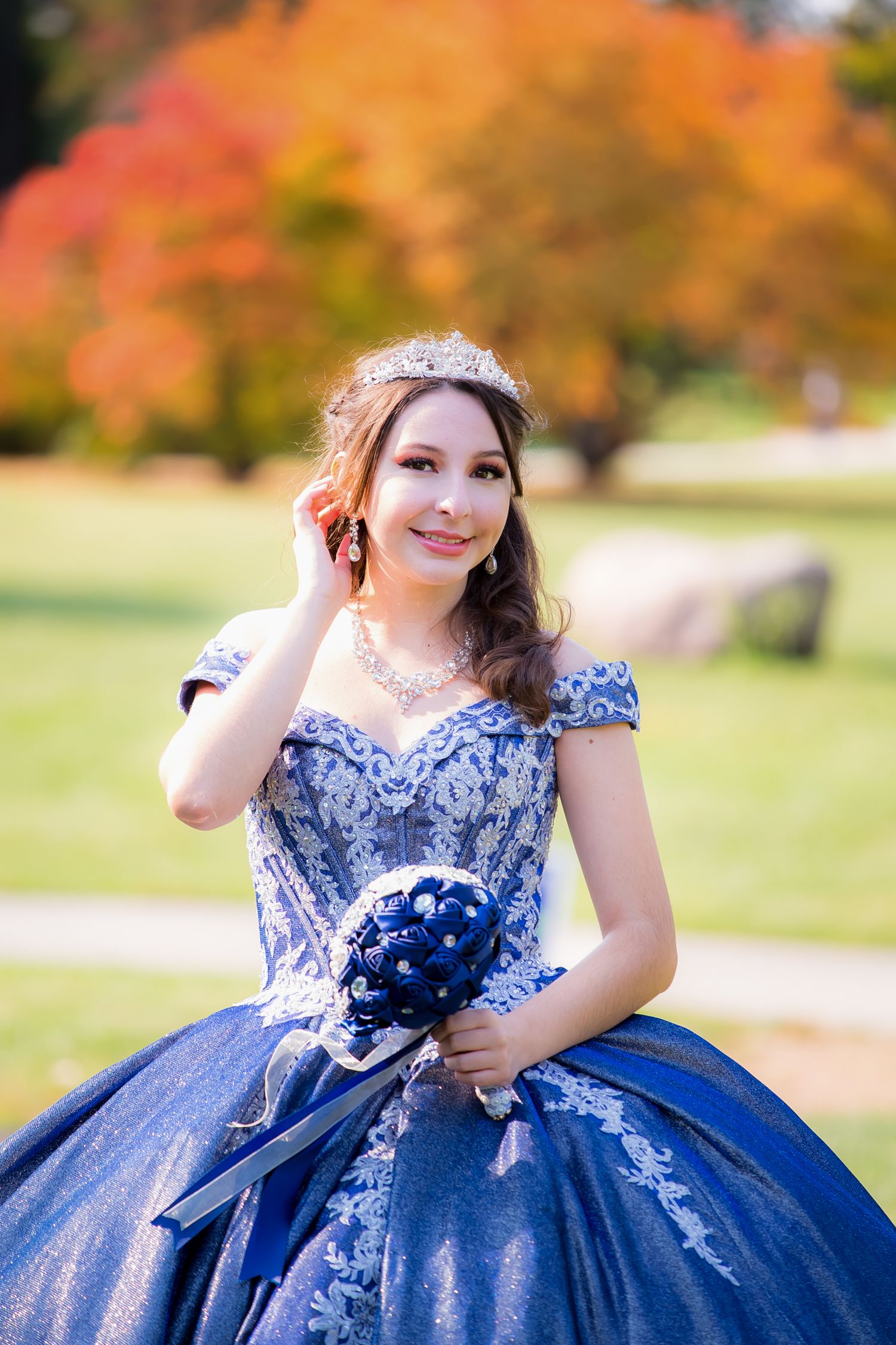 Young woman in blue ball gown and tiara poses outdoors during autumn.