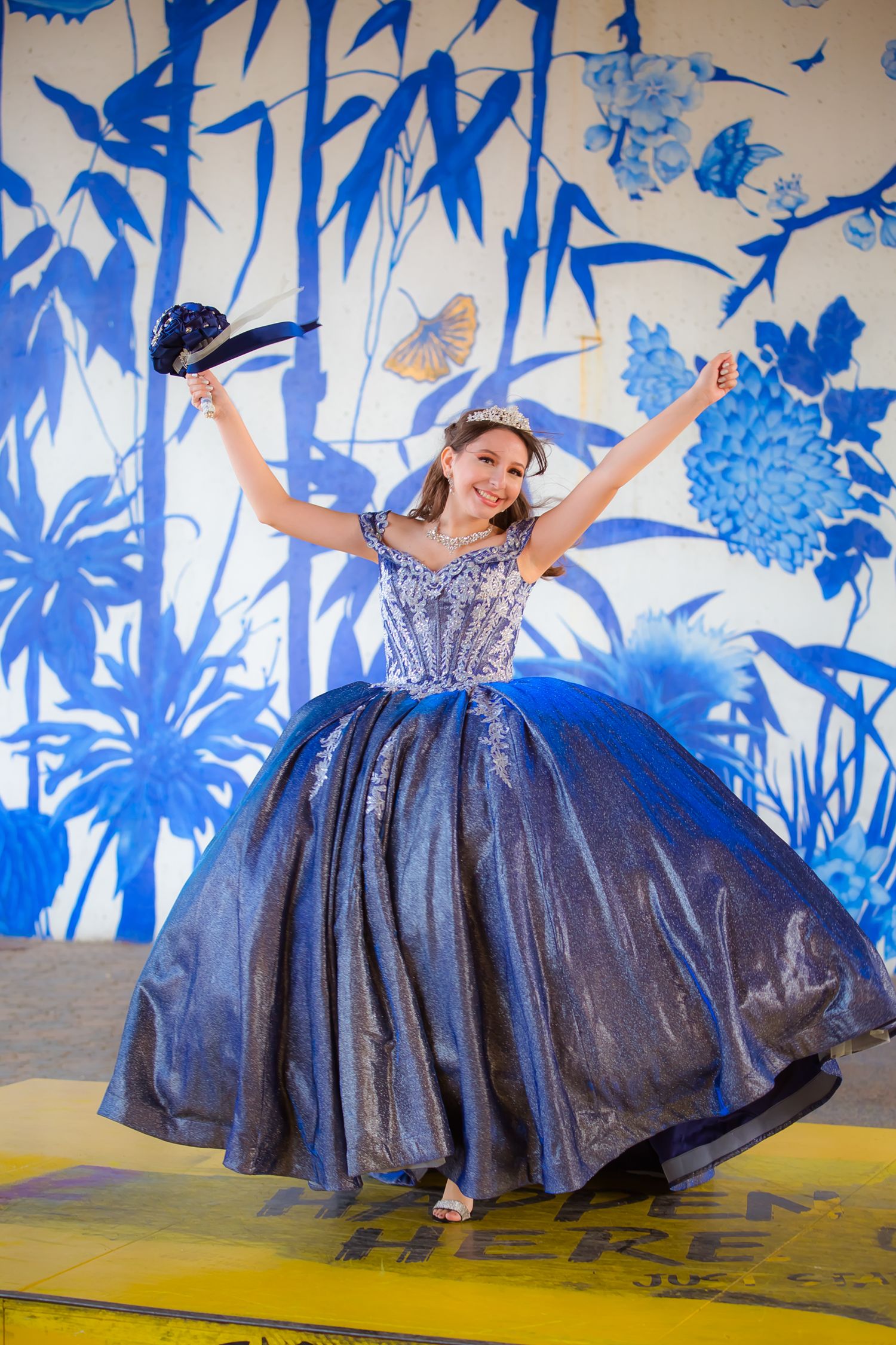 Joyful pose in a sparkly blue ball gown against blue floral wall art.