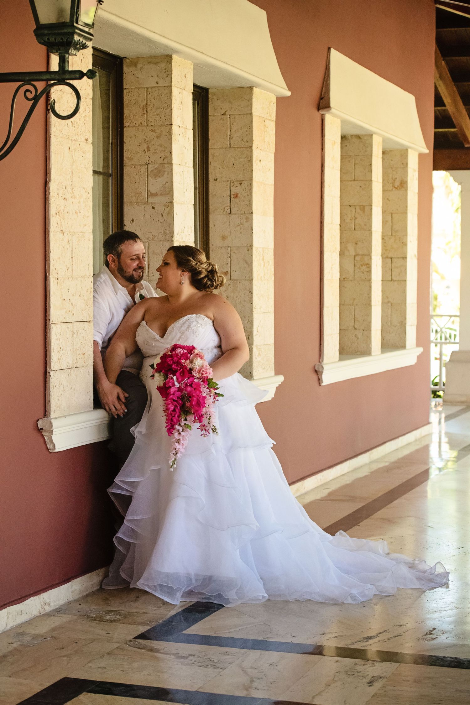 Bride in flowing white dress and groom sharing intimate moment in elegant hallway with coral walls.