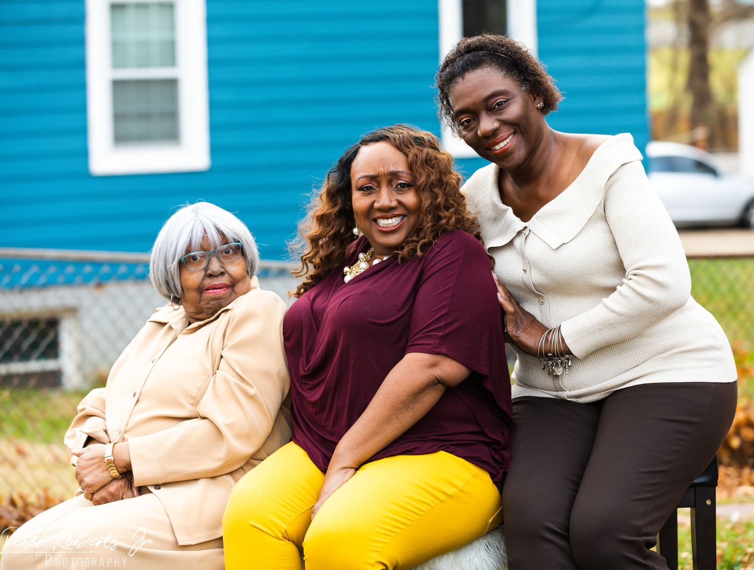Three smiling women pose together outdoors in front of a blue house for a family portrait.