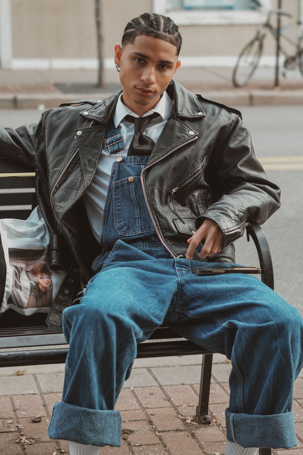 Casual model in denim overalls and leather jacket sitting on bench in urban setting.