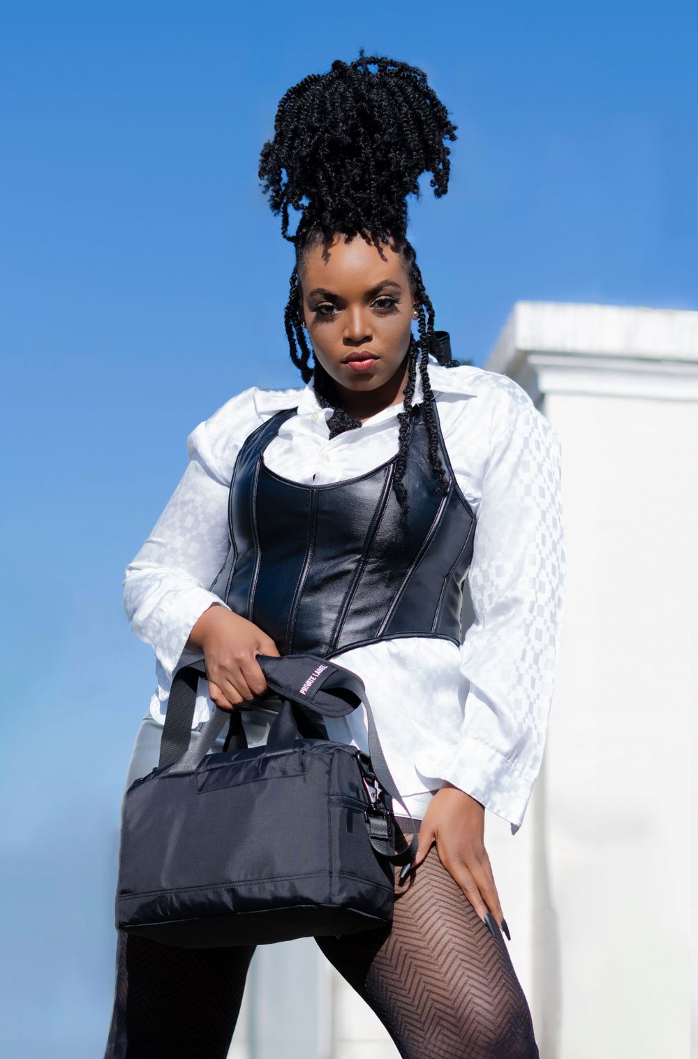 Fashion portrait of person in white shirt and black corset holding designer bag against blue sky.