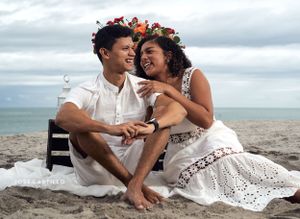 A romantic beach photo session with couple wearing flower crowns and white attire sitting on sand.