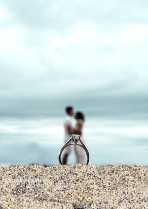 Close-up of engagement ring in sand with blurred silhouettes by ocean in background.