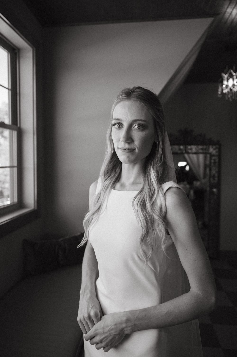Black and white portrait of someone in a minimalist white wedding dress by a window.