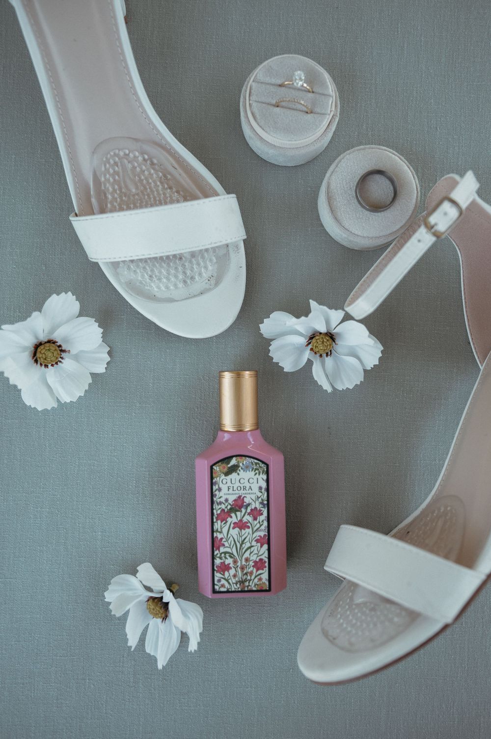 Bridal accessories including white sandals, perfume bottle and cosmos flowers on grey background.