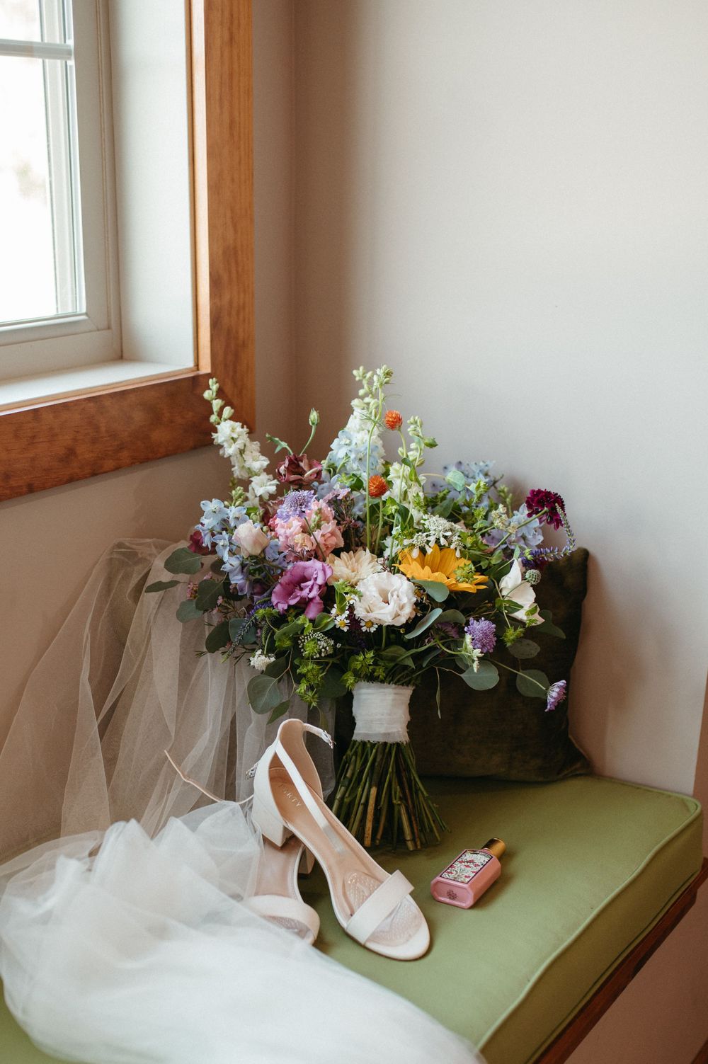 A bridal bouquet with purple and yellow flowers sits beside white shoes on a green cushioned bench.