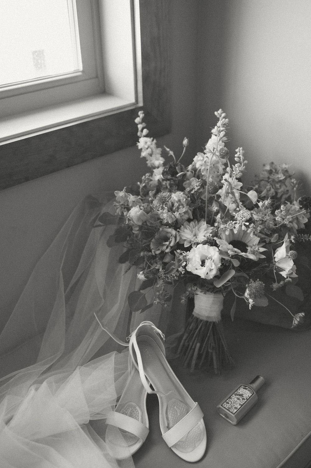 Black and white photo of wedding accessories including floral bouquet and bridal shoes near a window.