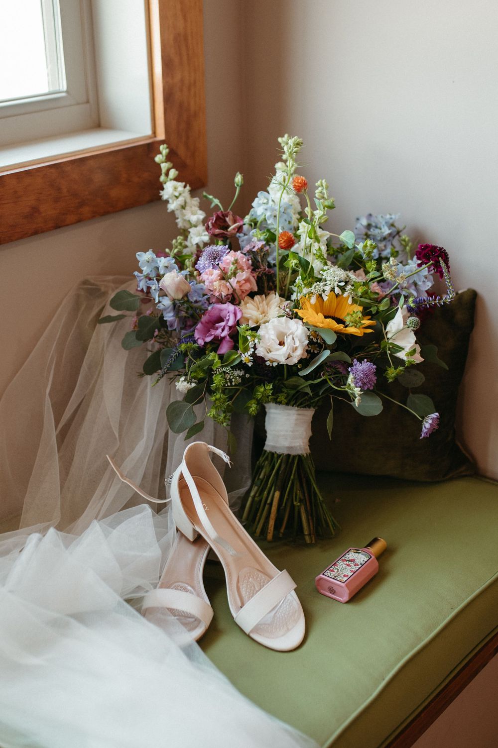Wedding details including a colorful wildflower bouquet, white heels, and veil arranged on a green bench.