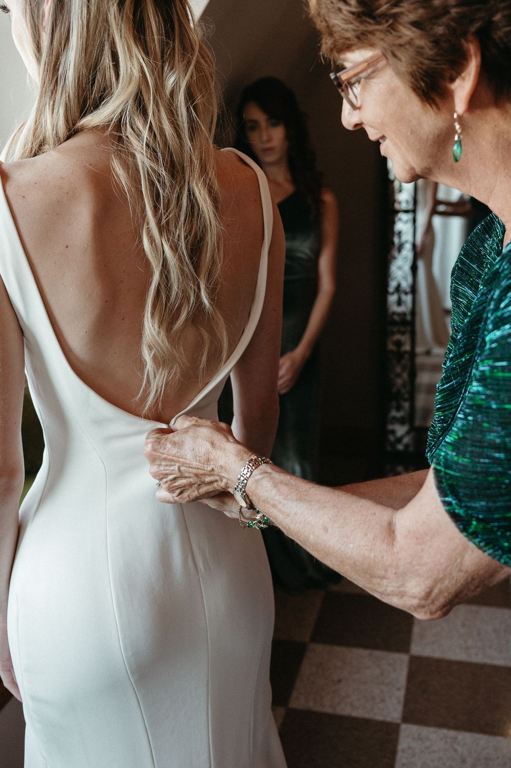 A woman in a white wedding dress gets final adjustments from someone wearing an emerald green dress.