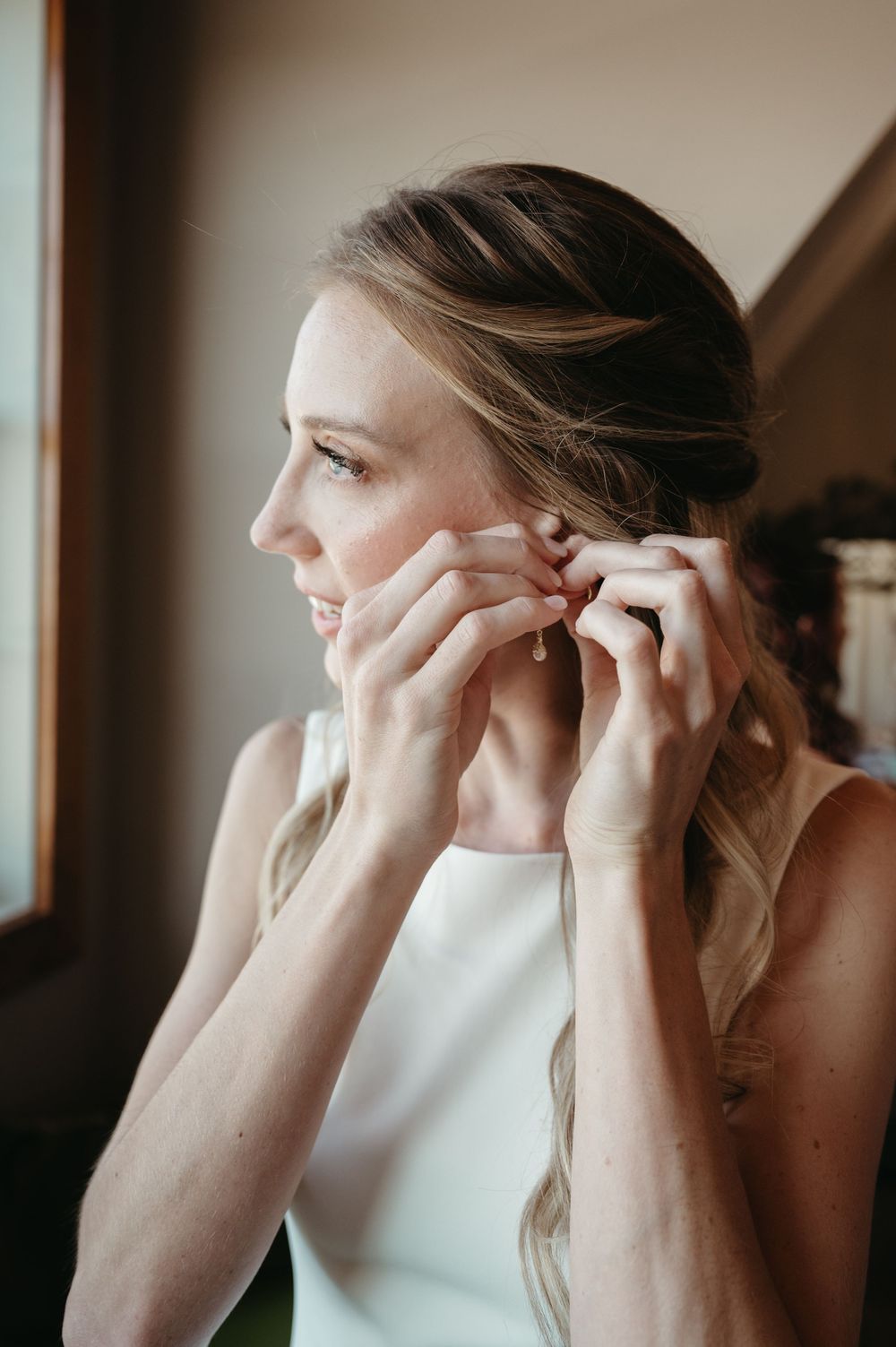 Close-up of someone putting on earrings while wearing a sleek white wedding dress.