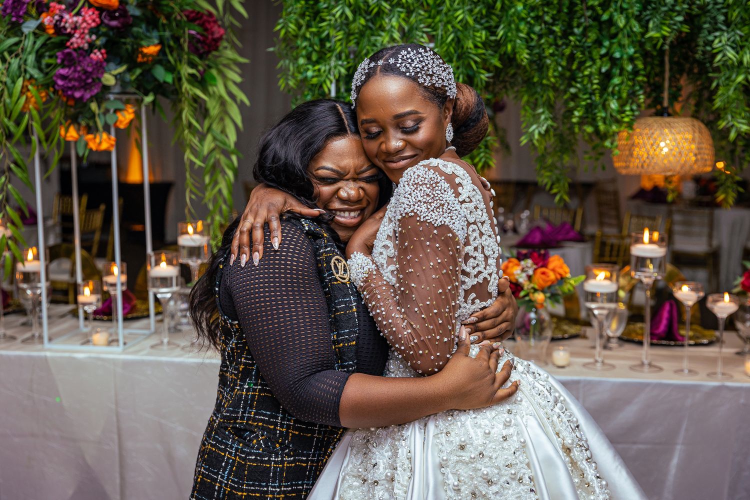 Two women in elegant attire embrace warmly at a festive event with decorative floral arrangements.