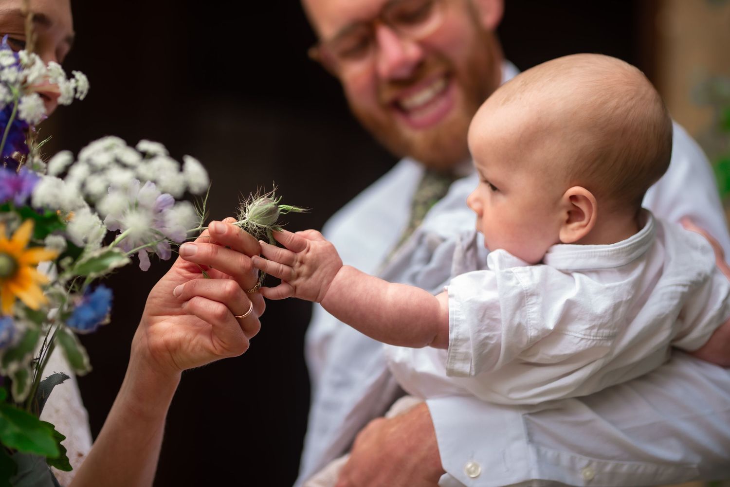baby in white reaches out to take a thistle from a wedding bouquet
