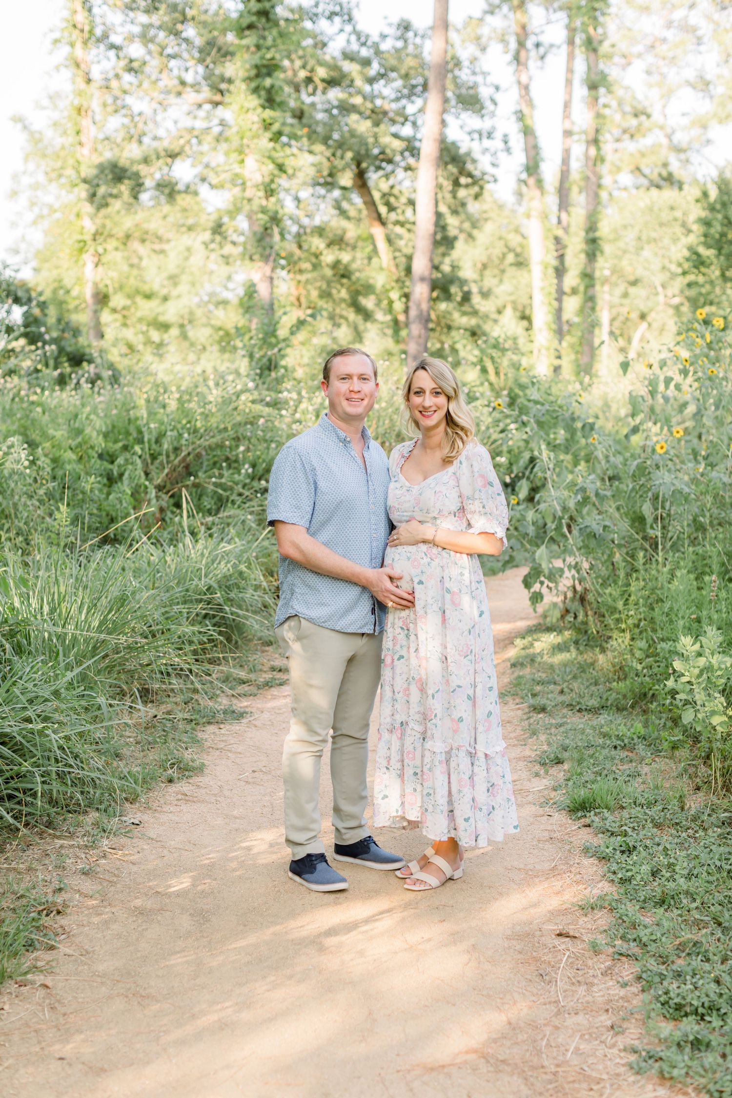 A couple stands together on a dirt path surrounded by lush greenery in a romantic maternity photo shoot.