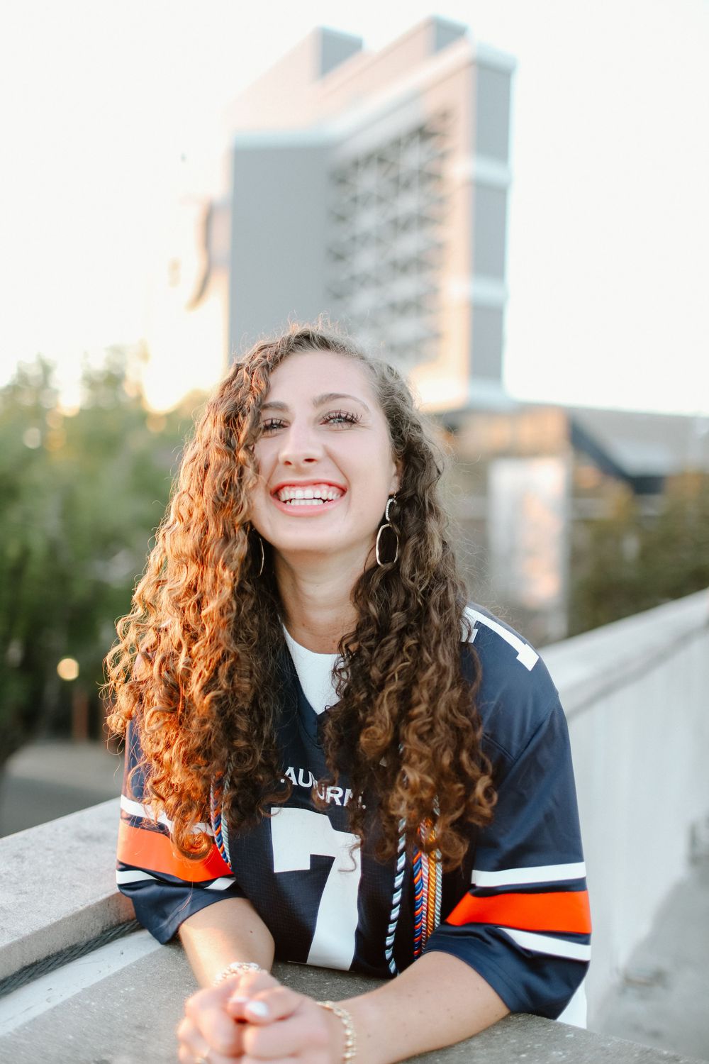 A person with curly hair wearing a navy and orange athletic jersey smiles brightly in an outdoor setting.