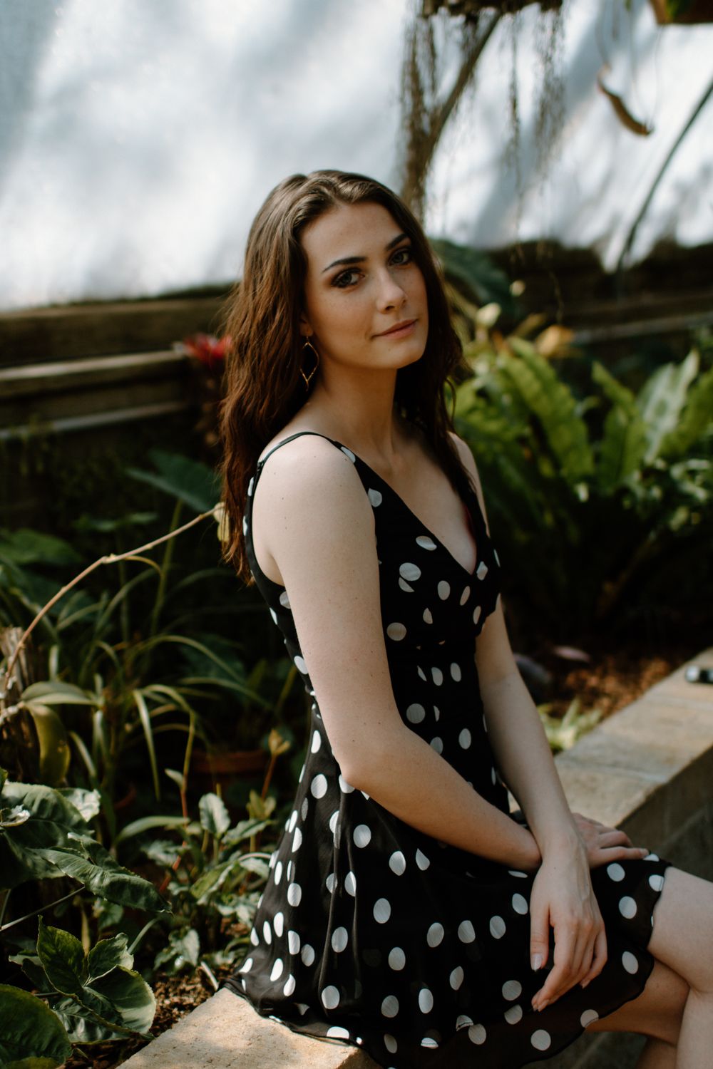 A person in a polka dot dress poses outdoors in a garden setting with natural lighting and greenery in the background.
