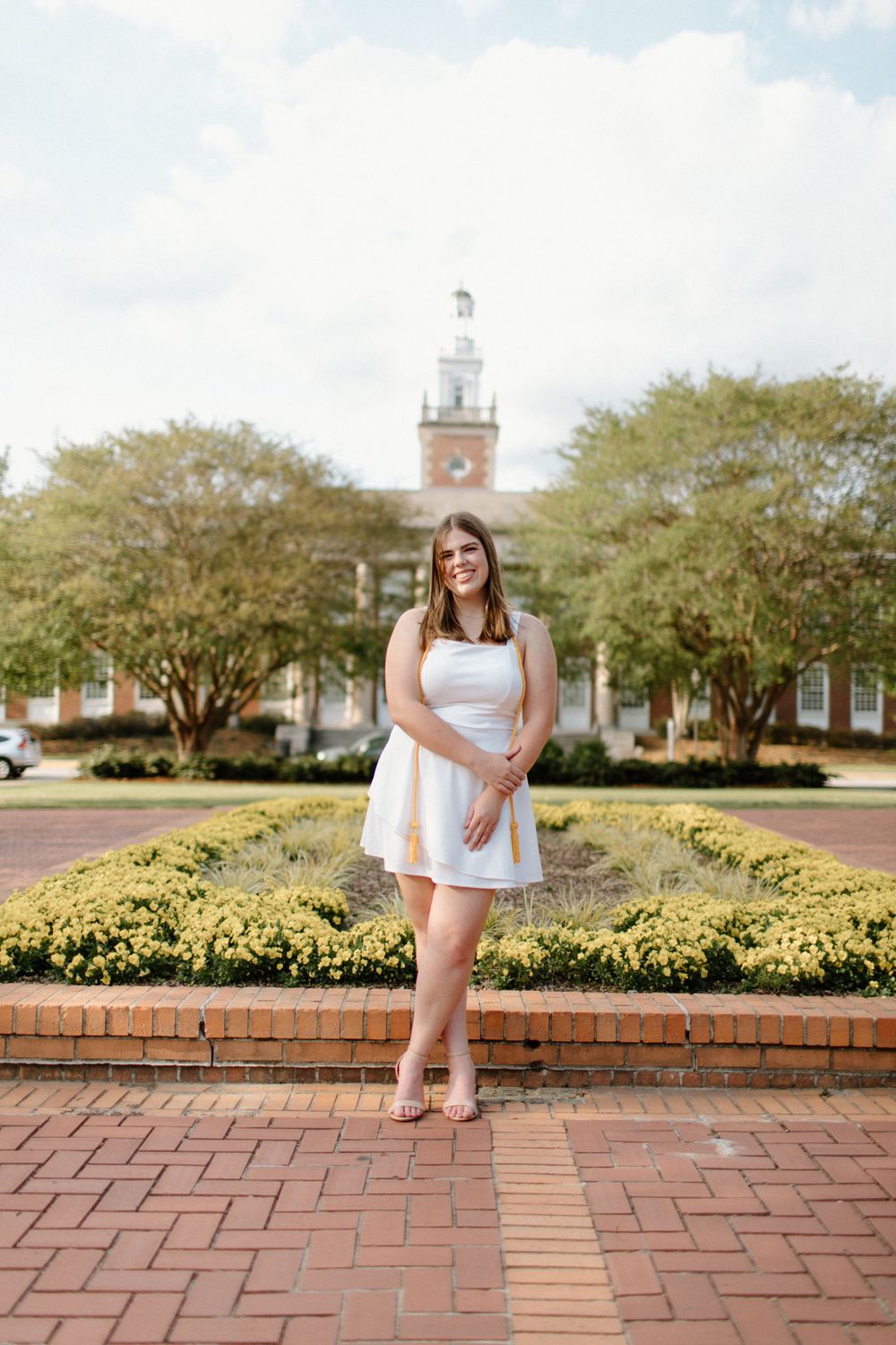 A white dress-clad figure stands on brick pavers in front of a landscaped garden and historic building with a clock tower.