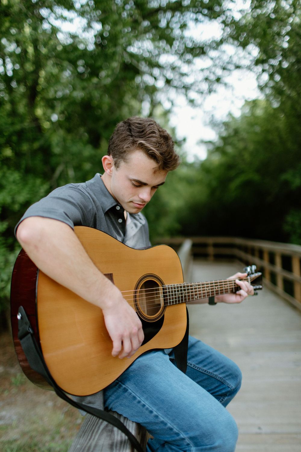 Musician in jeans plays acoustic guitar while sitting on wooden boardwalk in natural outdoor setting.