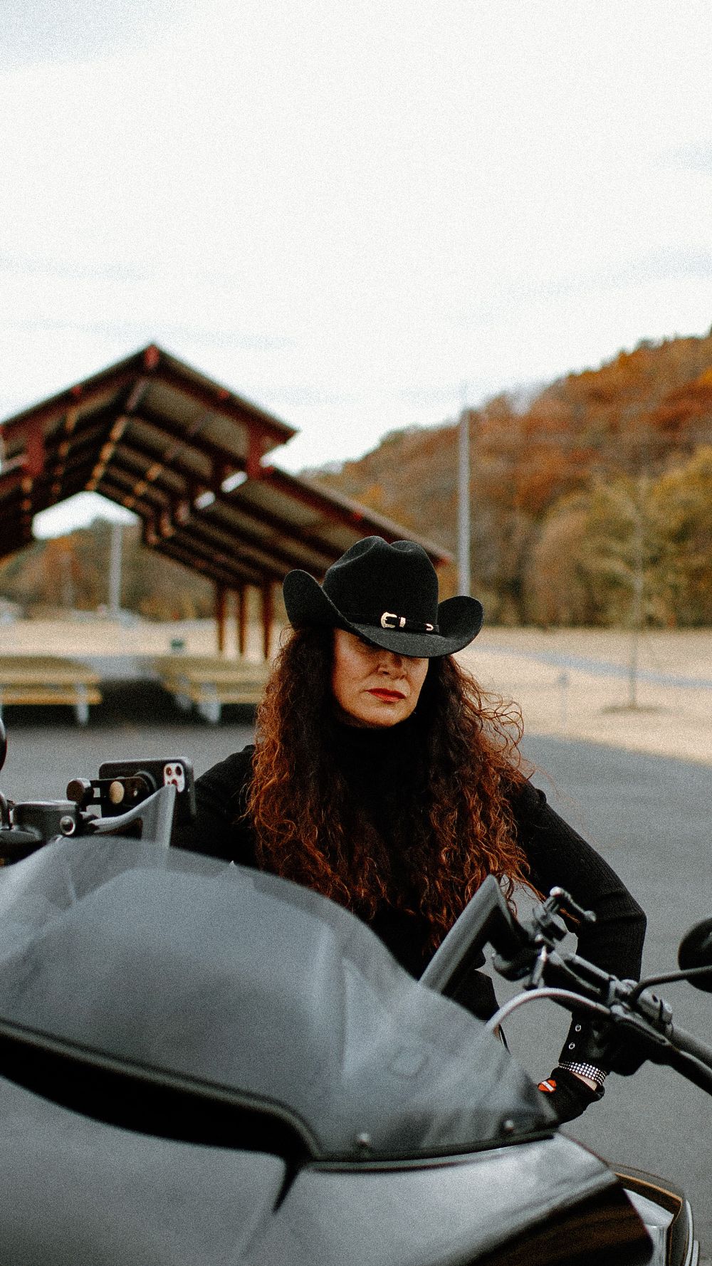 A motorcyclist in a black cowboy hat and long hair sits on a parked motorcycle on a cloudy day.