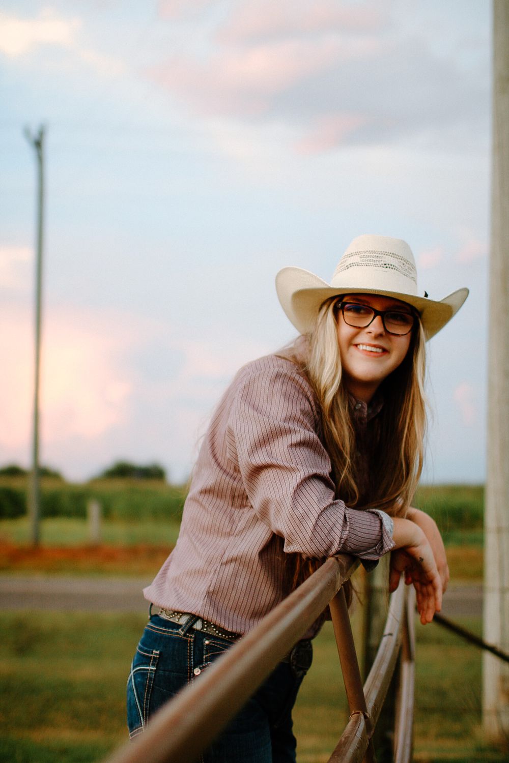 A person in a cowboy hat and striped shirt leans on a rustic fence post at sunset in a rural field.