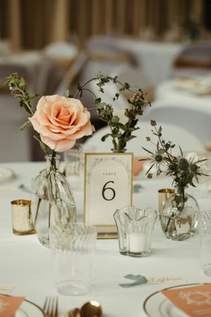 Wedding reception table settings with delicate peach roses in small glass vases and gold table numbers.