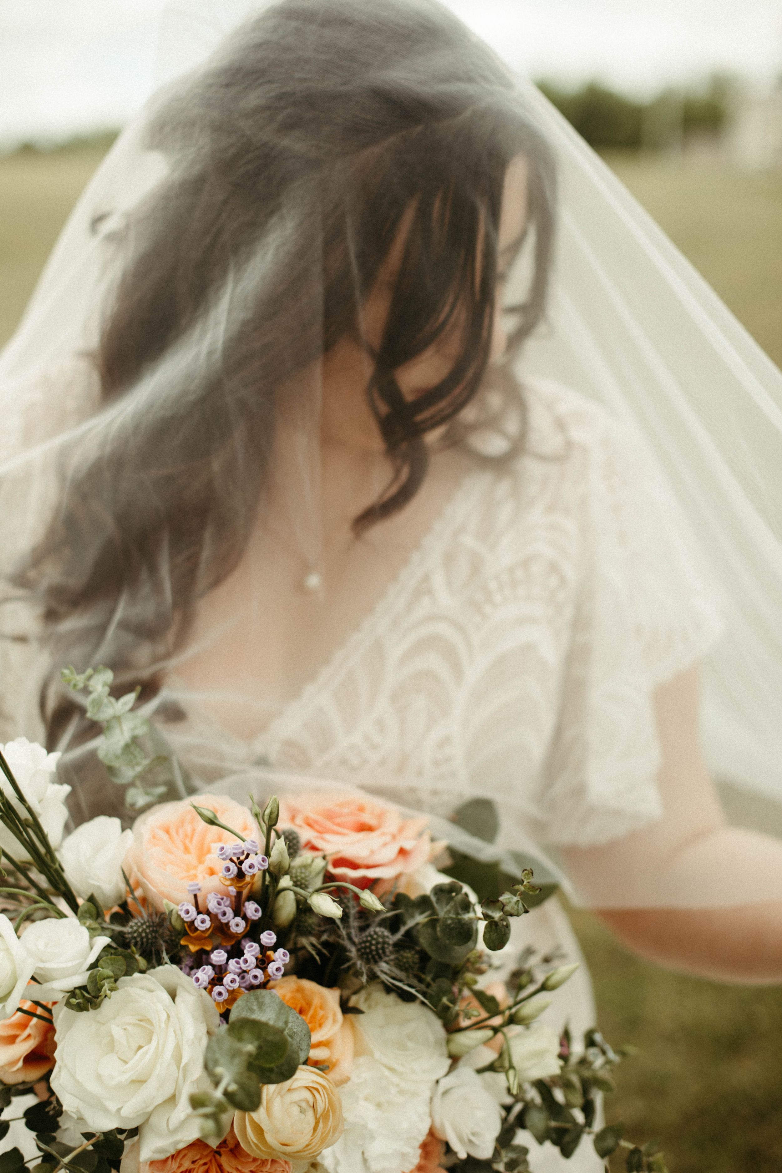 Bridal bouquet featuring peach and white roses with eucalyptus leaves against a flowing veil.