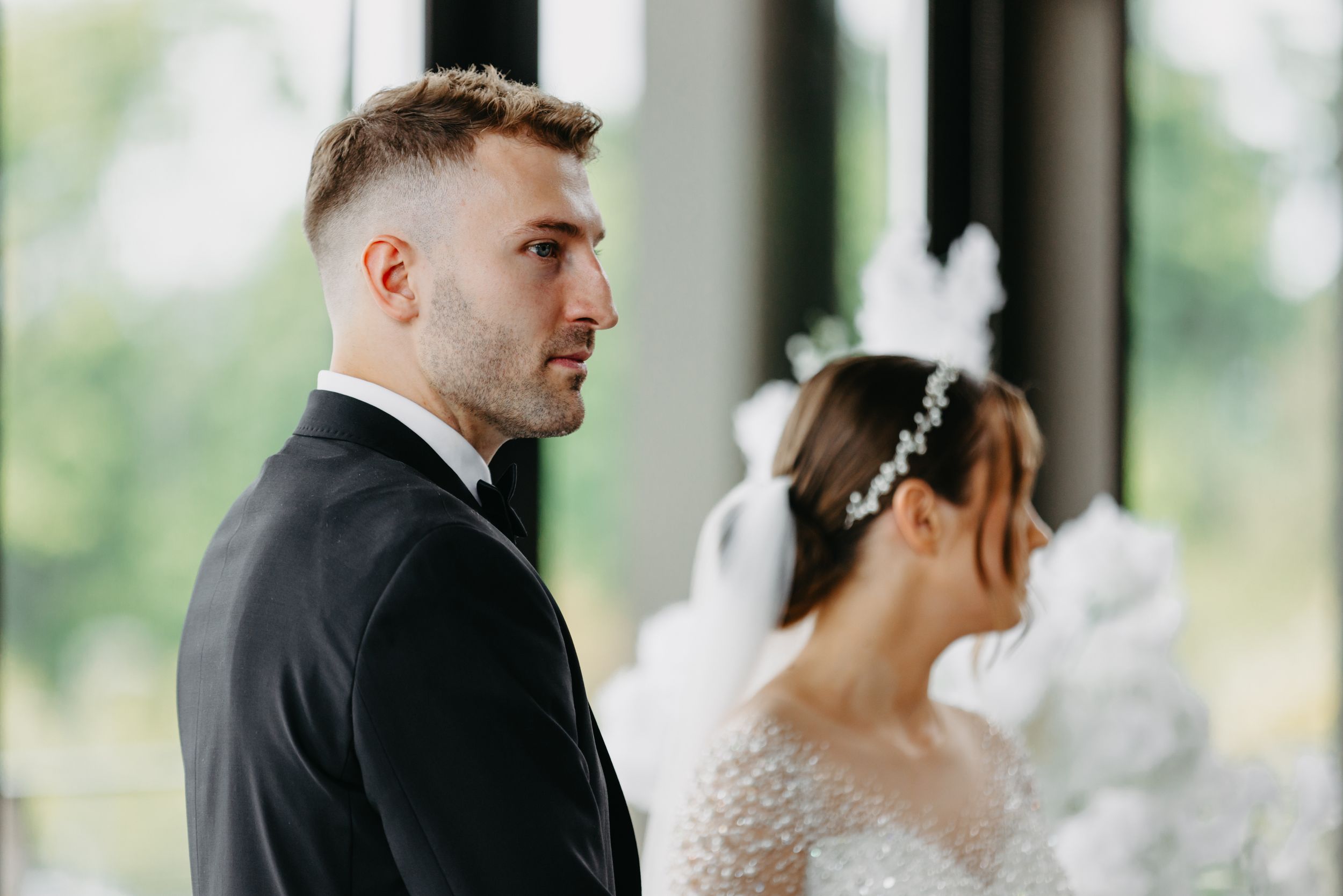 A couple in formal wedding attire stands in an elegant venue with large windows.