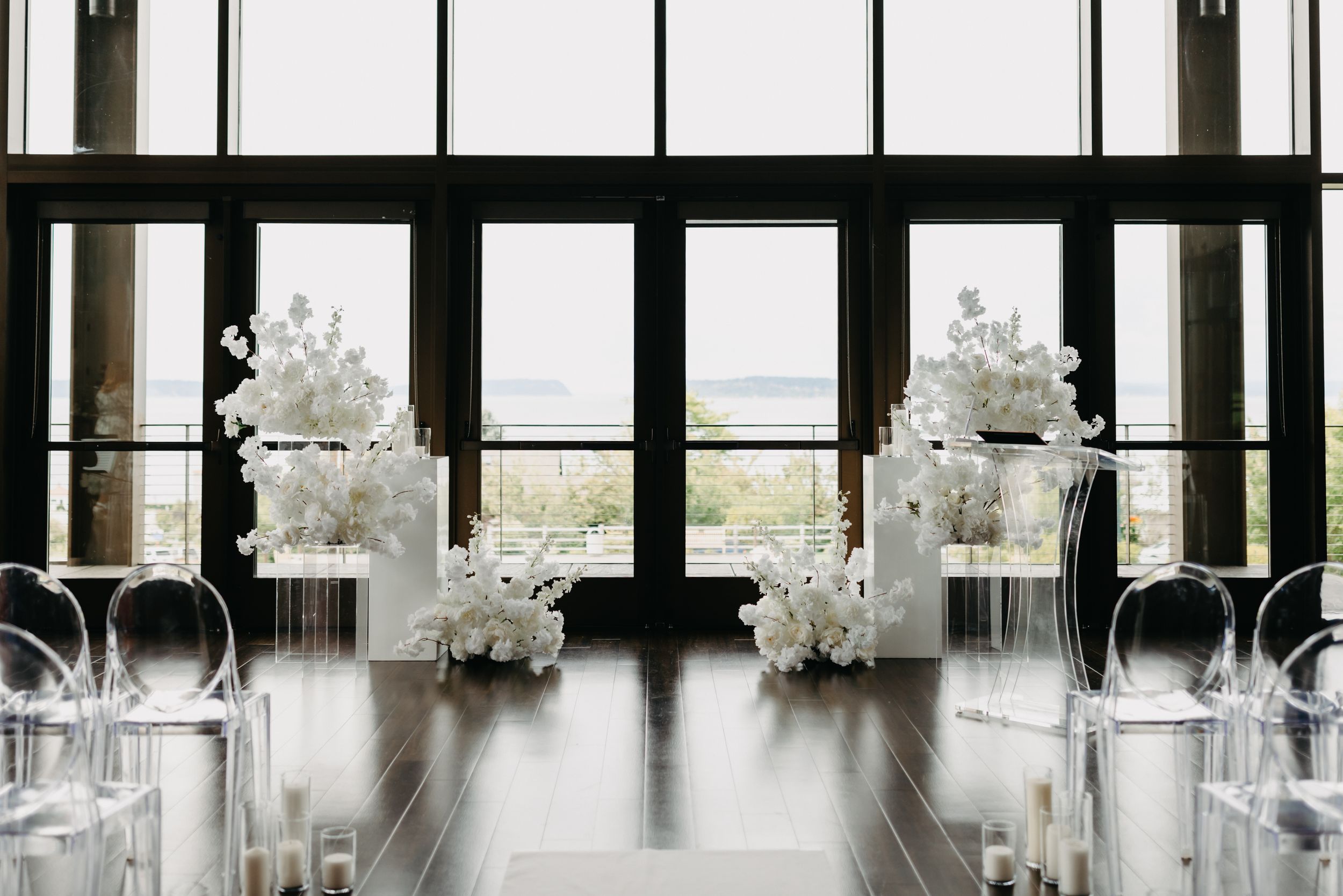 Modern wedding venue setup with ghost chairs and white floral arrangements against large windows.