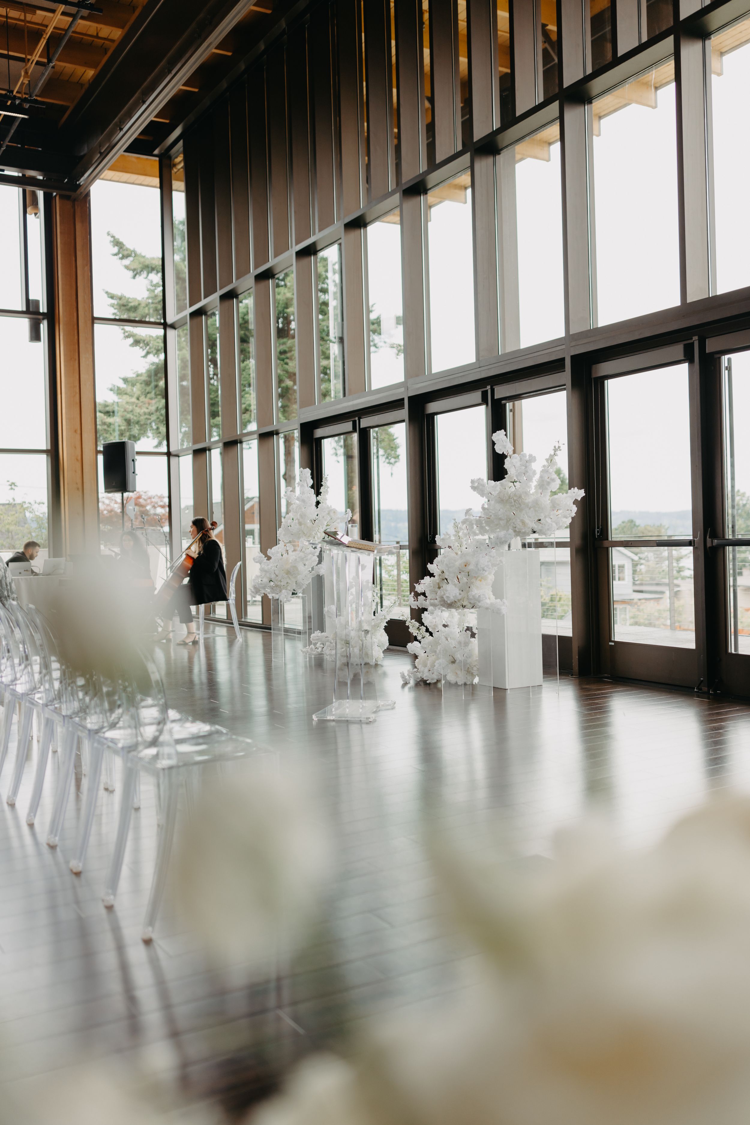 Modern event venue with white floral arrangements and ghost chairs along floor-to-ceiling windows.