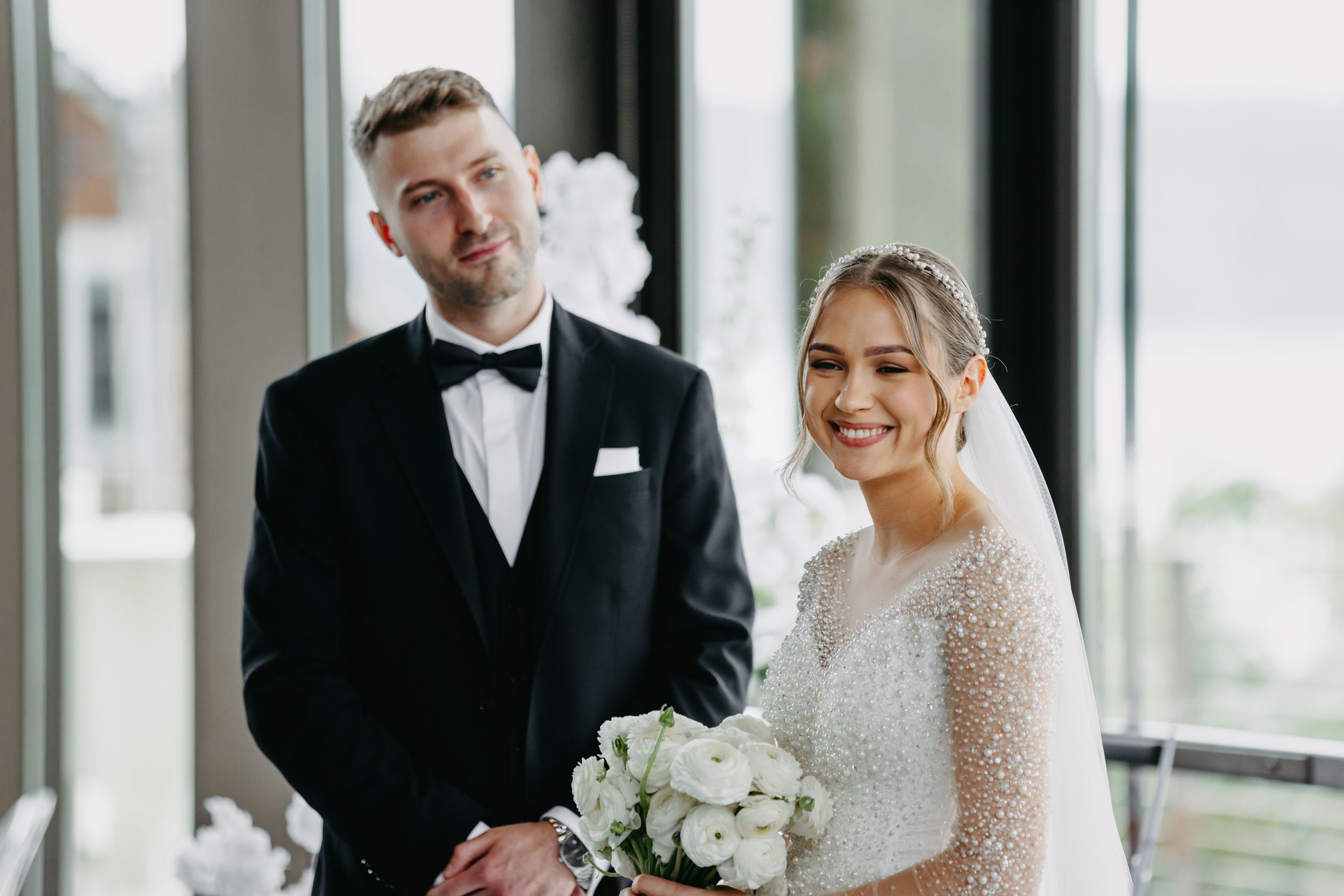 A couple in wedding attire smiles together in front of large windows.