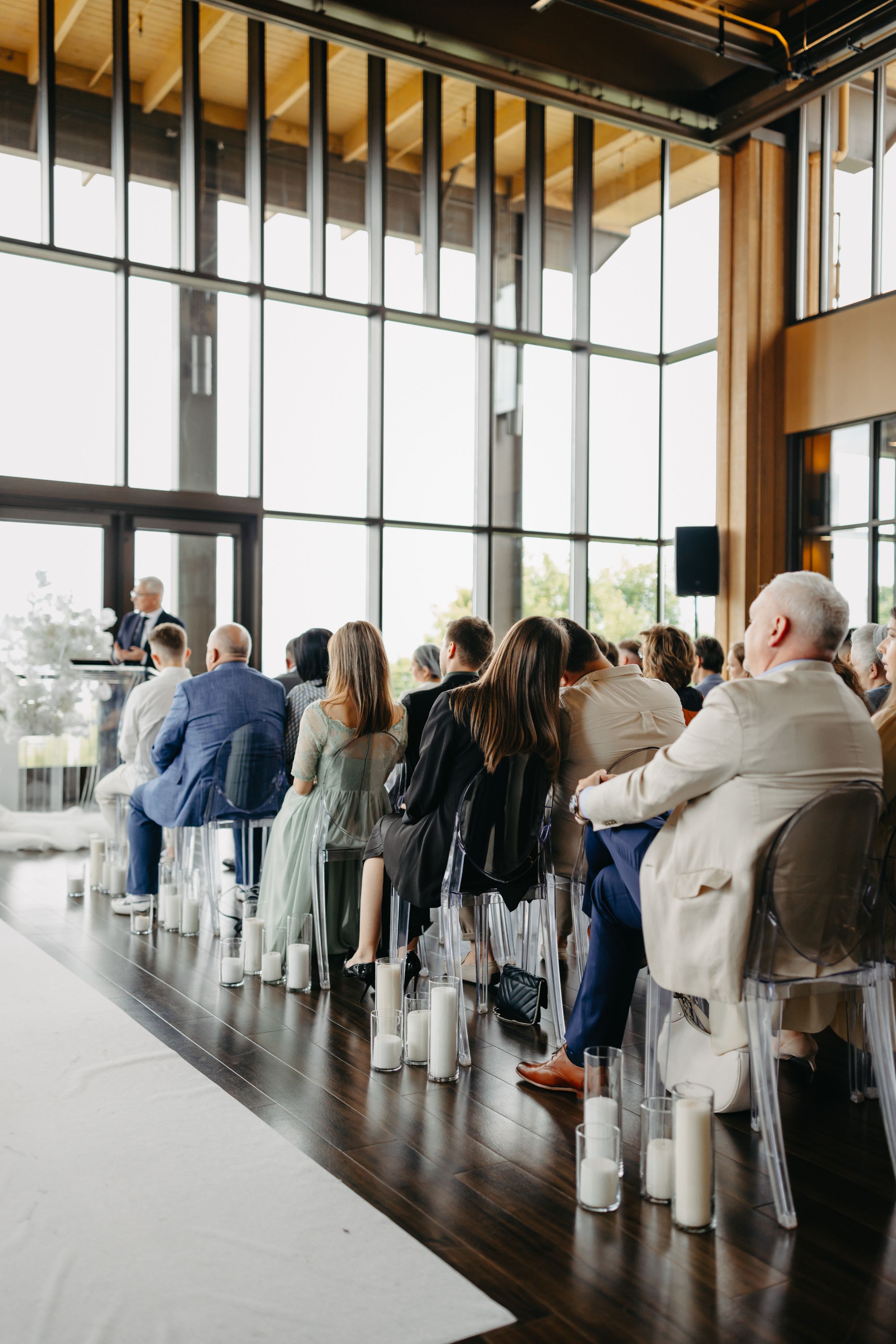 Wedding guests seated in rows at an elegant venue with large windows and natural light.
