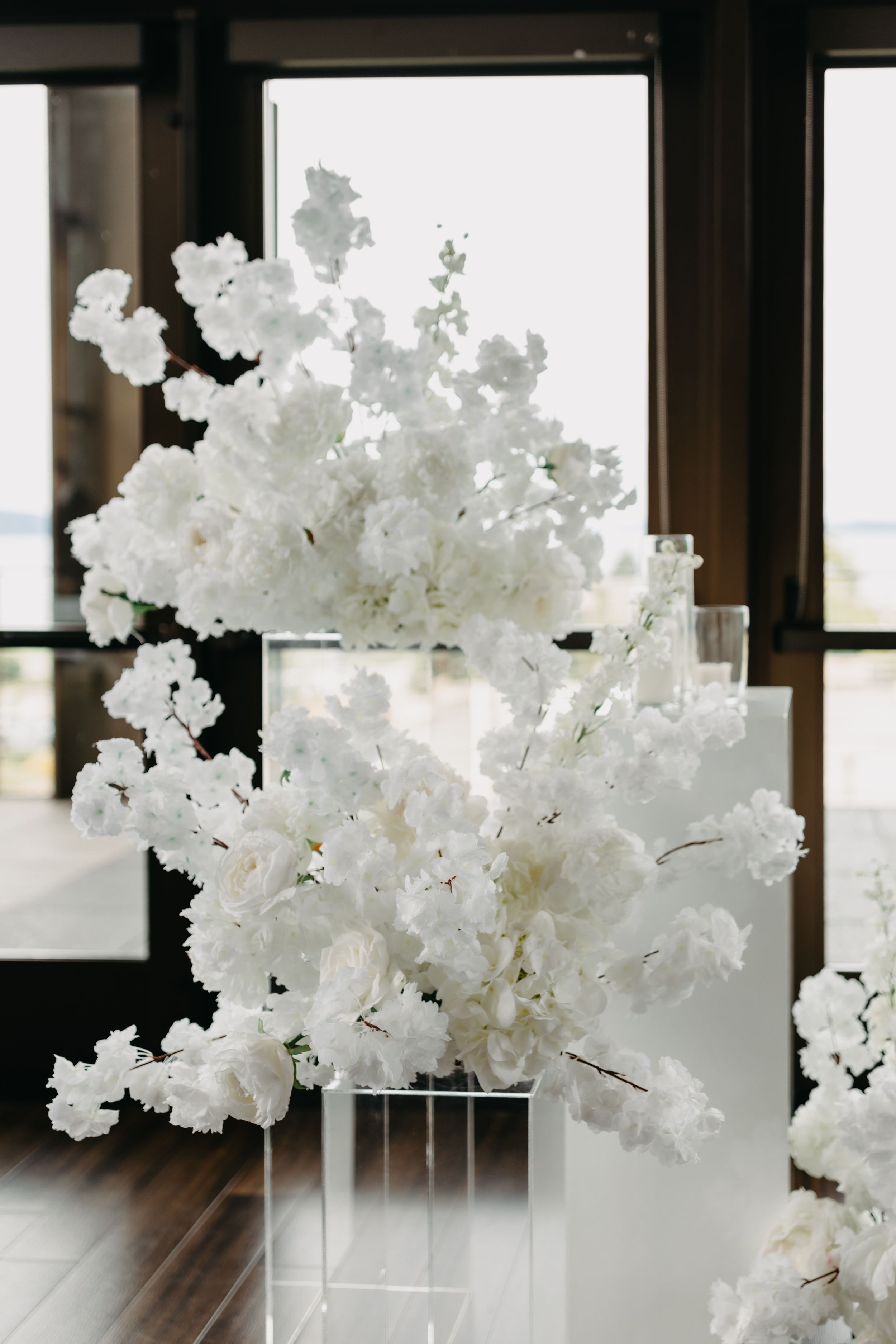 Elegant white floral arrangement on clear acrylic pedestal with window backdrop.