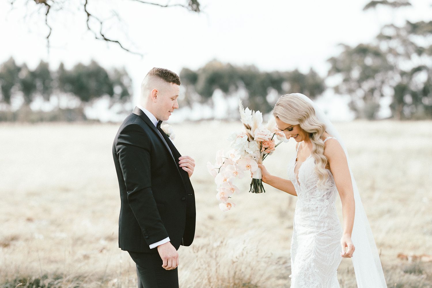 A couple in wedding attire share a joyful moment in an outdoor field with a soft, dreamy background.
