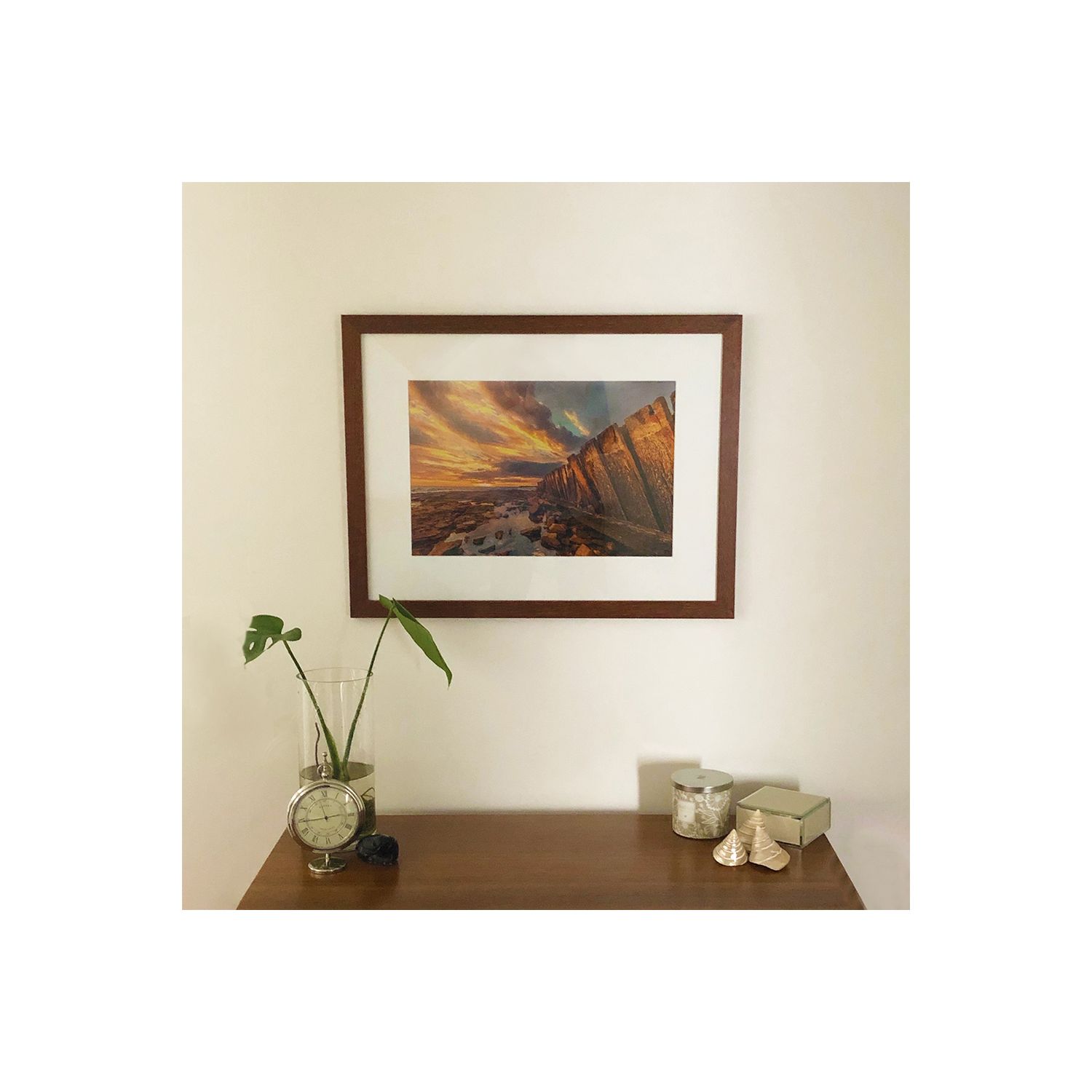 Brown framed landscape photograph of mountains at sunset displayed above wooden table with decorative items.