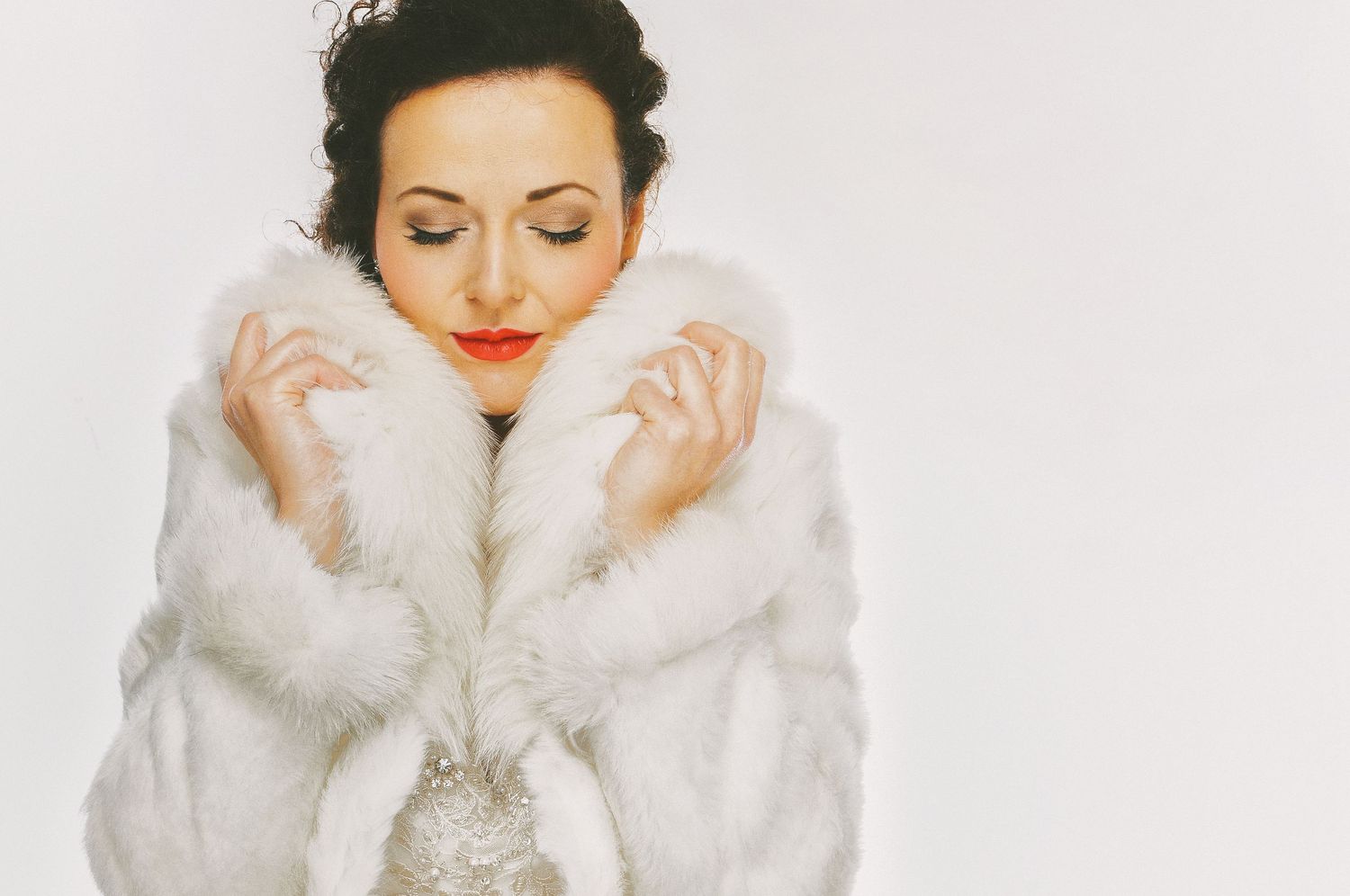 A luxurious white fur coat worn with closed eyes and red lipstick in a soft, vintage-style portrait.