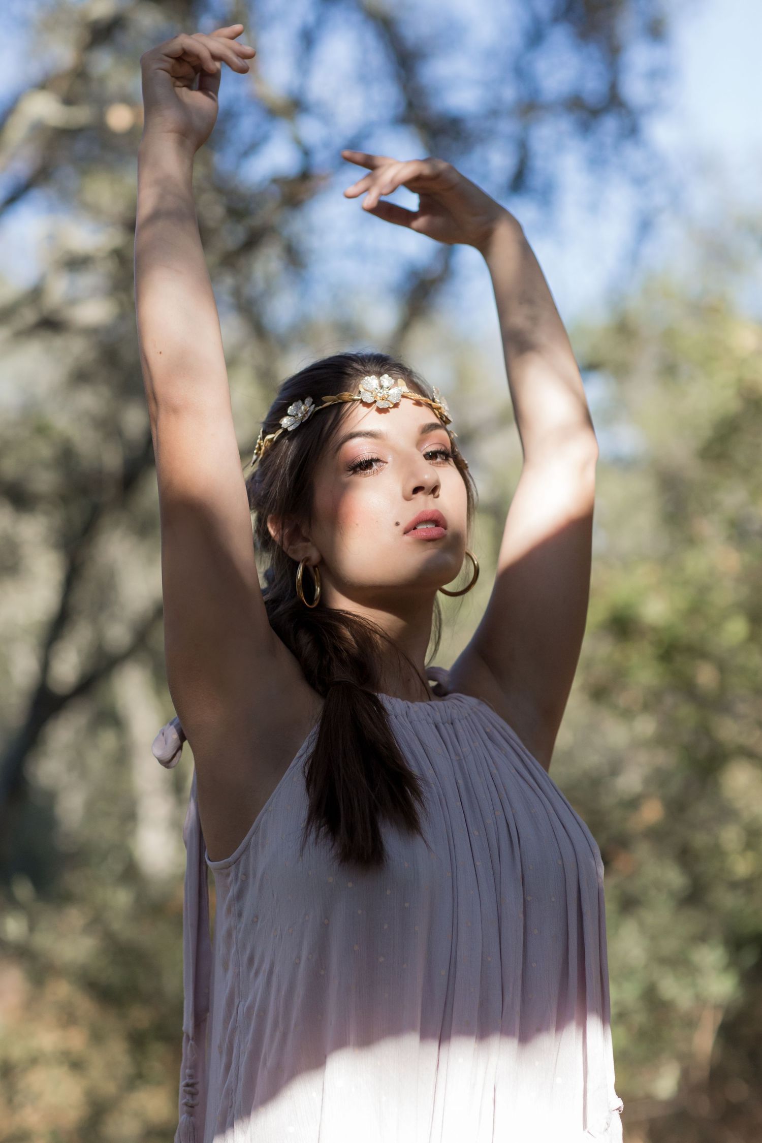 Young woman in lavender dress raising arms gracefully while wearing a gold floral headband outdoors.