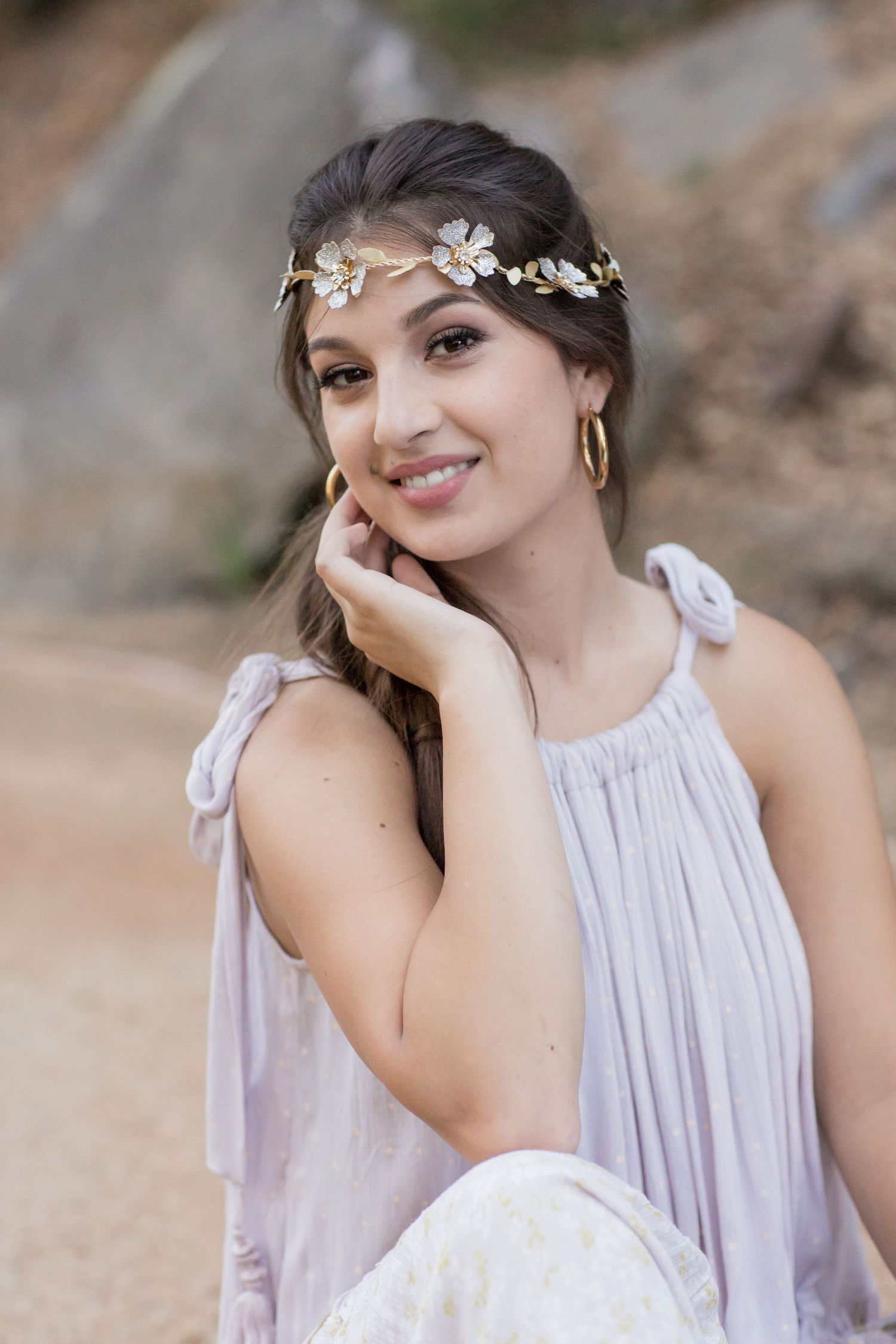 Young woman in lavender top with gold floral headpiece smiling softly while touching her cheek.