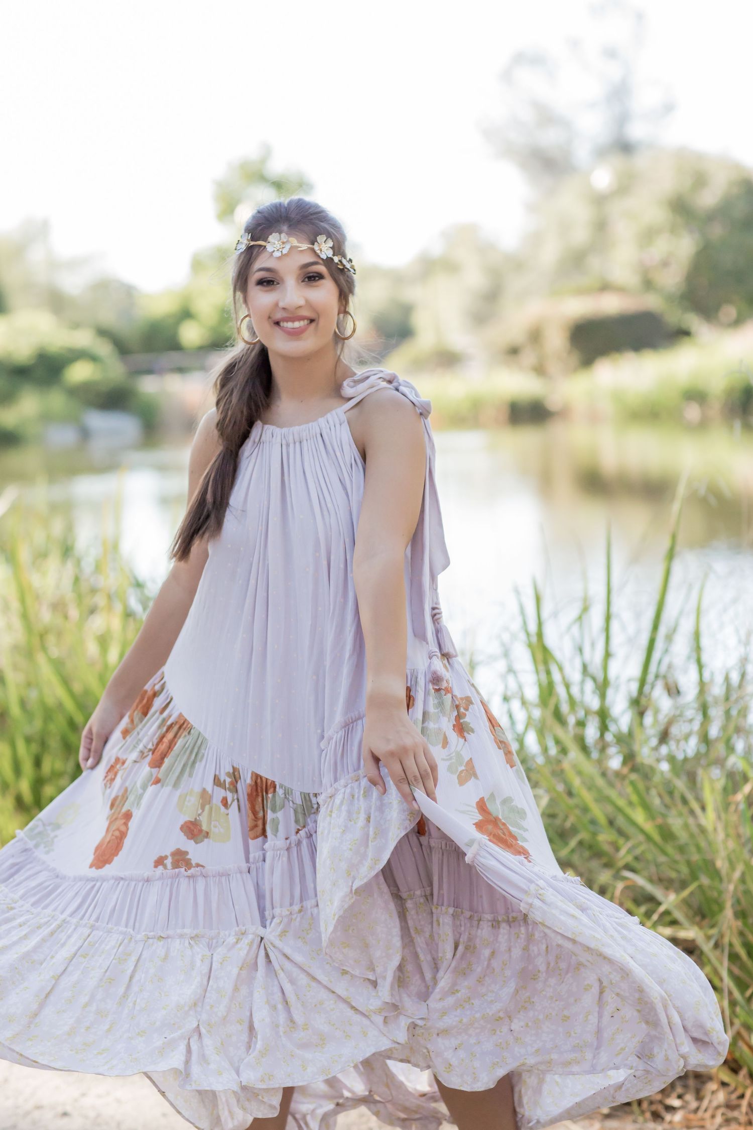 Young woman in flowy lavender boho maxi dress and floral crown twirling outdoors near a pond.