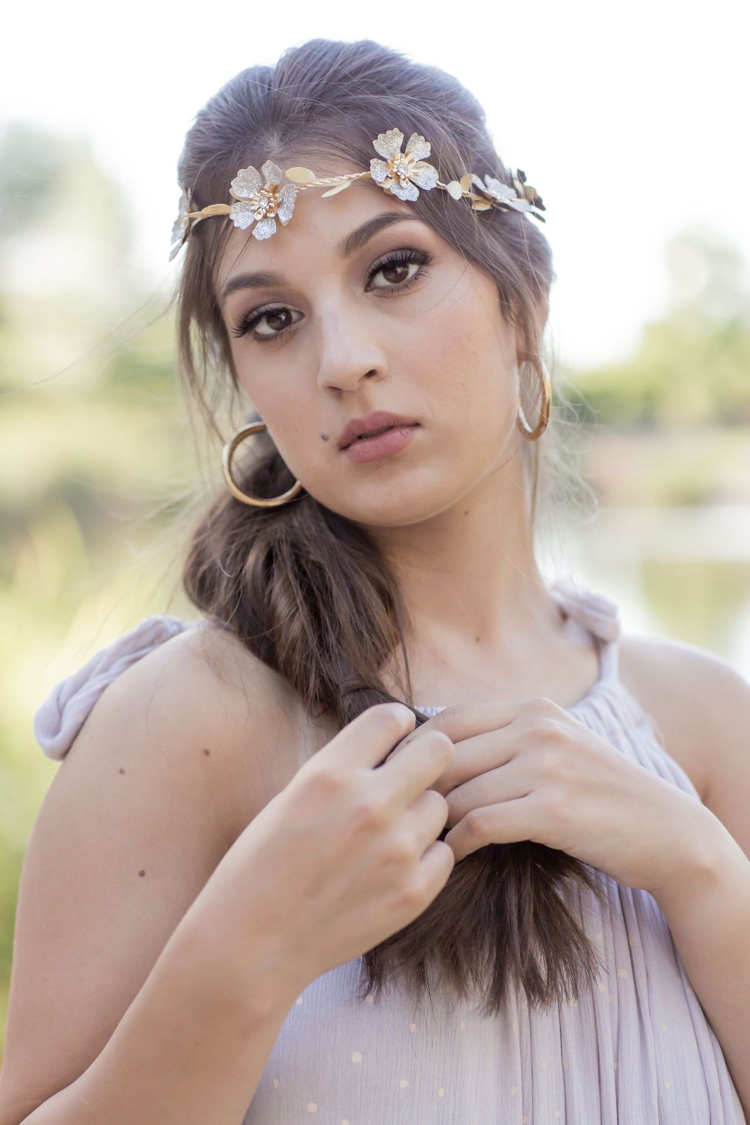 Close-up of young woman wearing gold floral crown and hoop earrings in soft lavender dress outdoors.