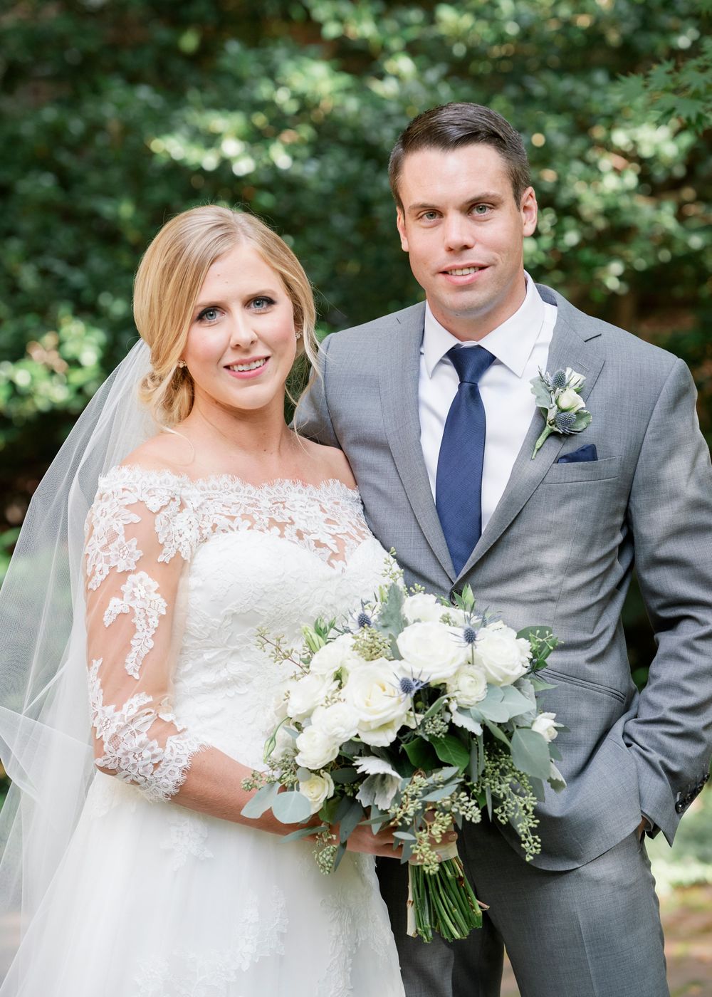 Wedding couple outside poses together with bride holding elegant white and green floral bouquet in garden setting.