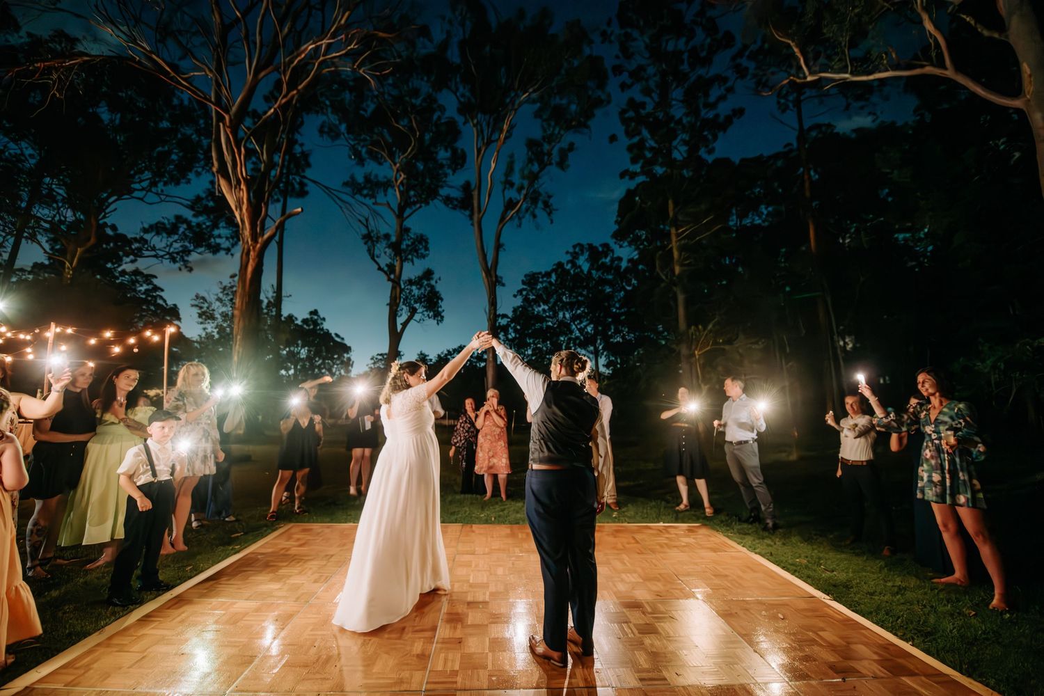 Newlyweds dancing on illuminated outdoor dance floor surrounded by string lights and tall trees.