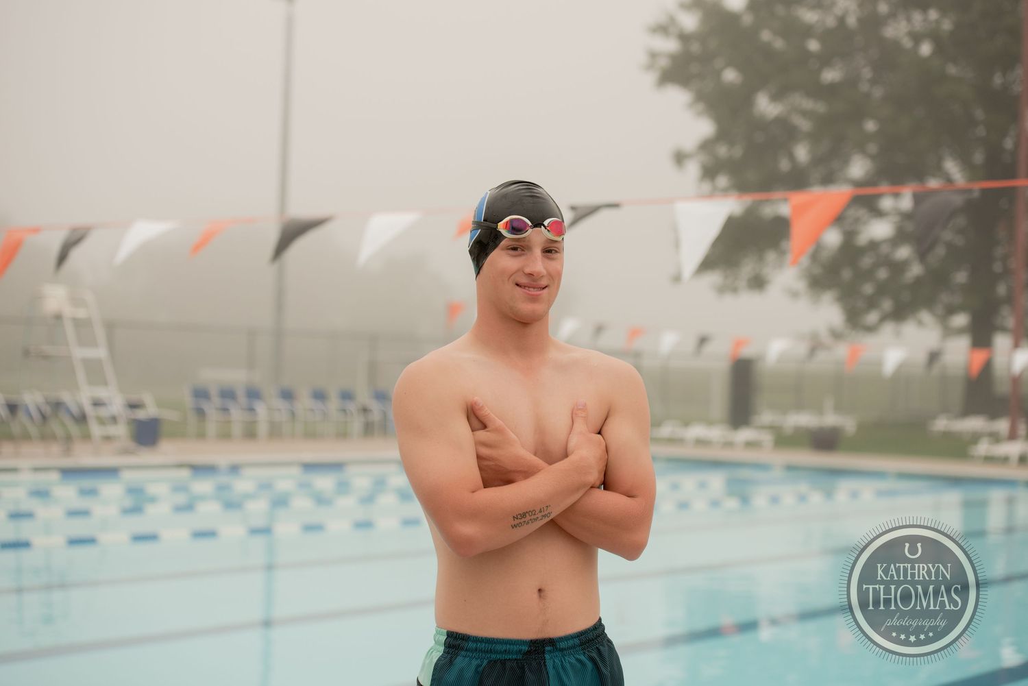 A swimmer in swim cap and goggles stands by pool with decorative flags on foggy day.