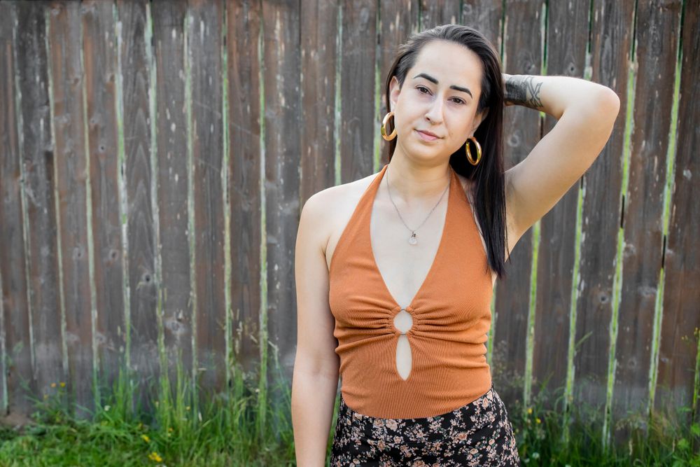 Model in orange halter top and floral skirt poses casually against wooden fence.