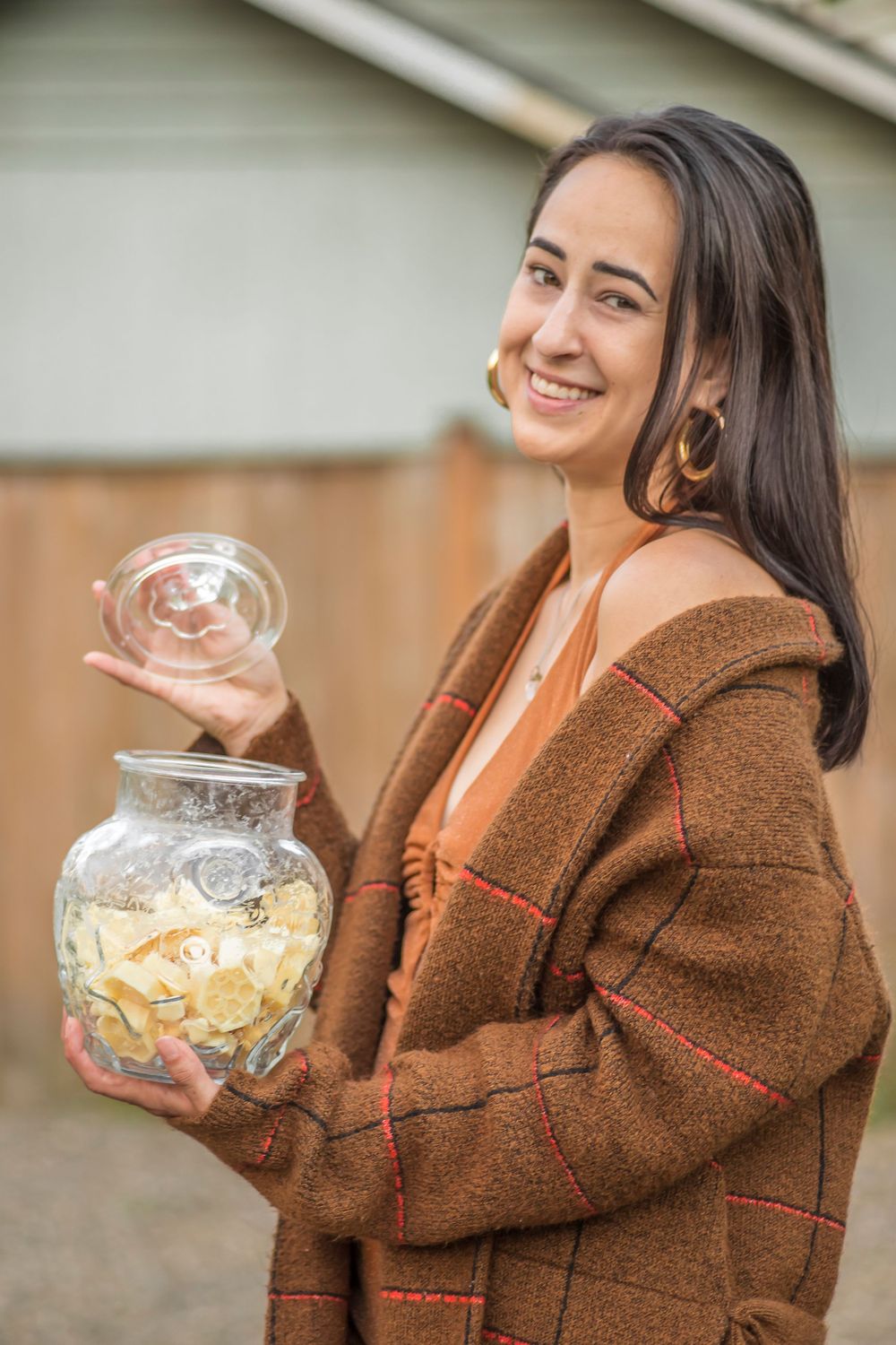 Woman in brown cardigan holds glass jar filled with snacks while smiling outdoors.