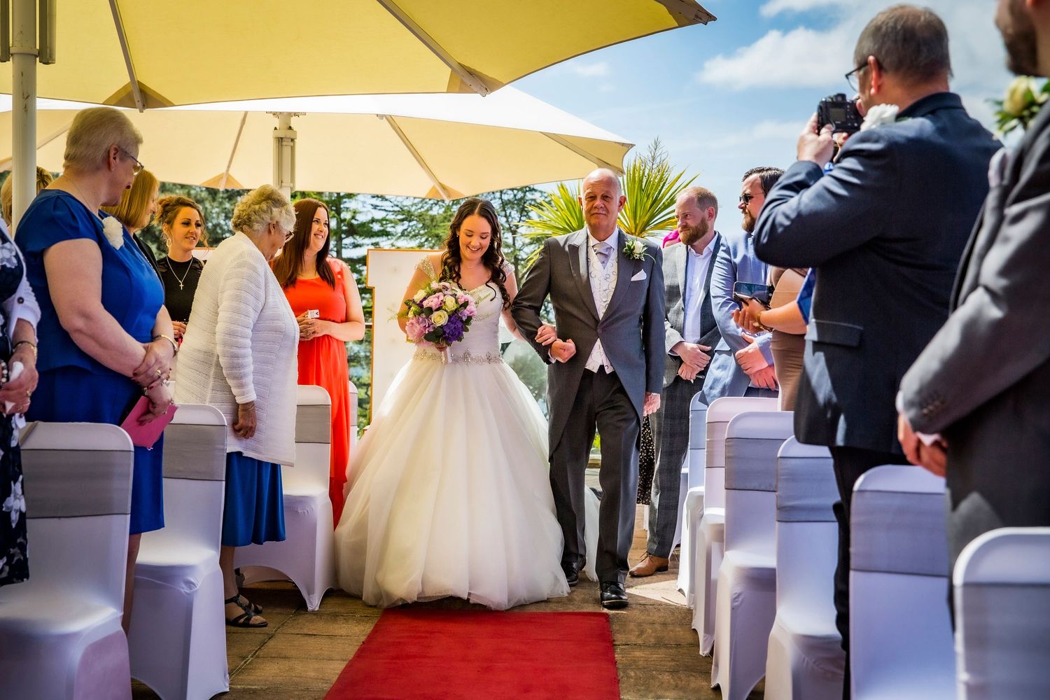 An outdoor wedding ceremony under yellow umbrellas with guests seated on white chairs.